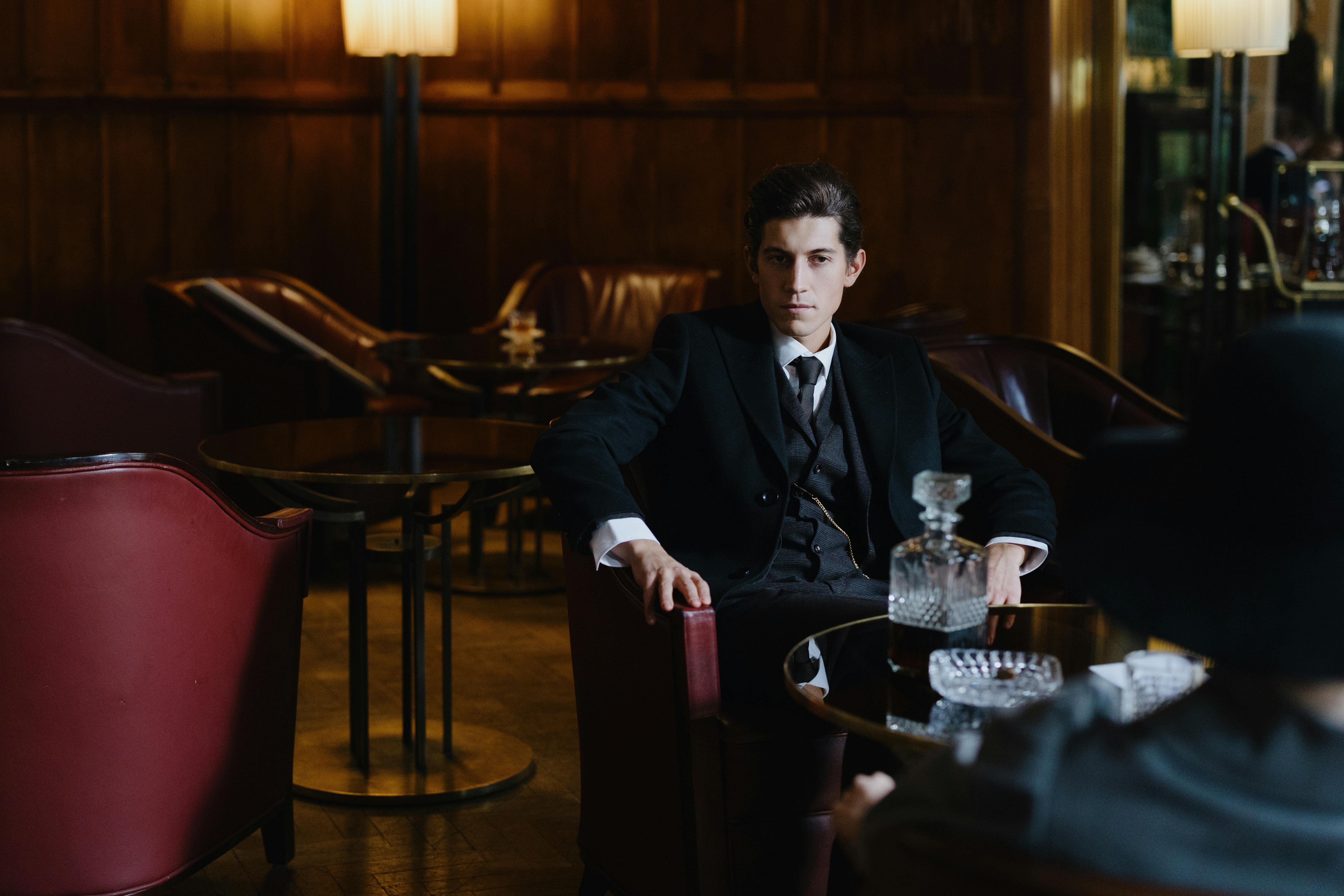 Man in suit sits in a dimly lit lounge with decanter.