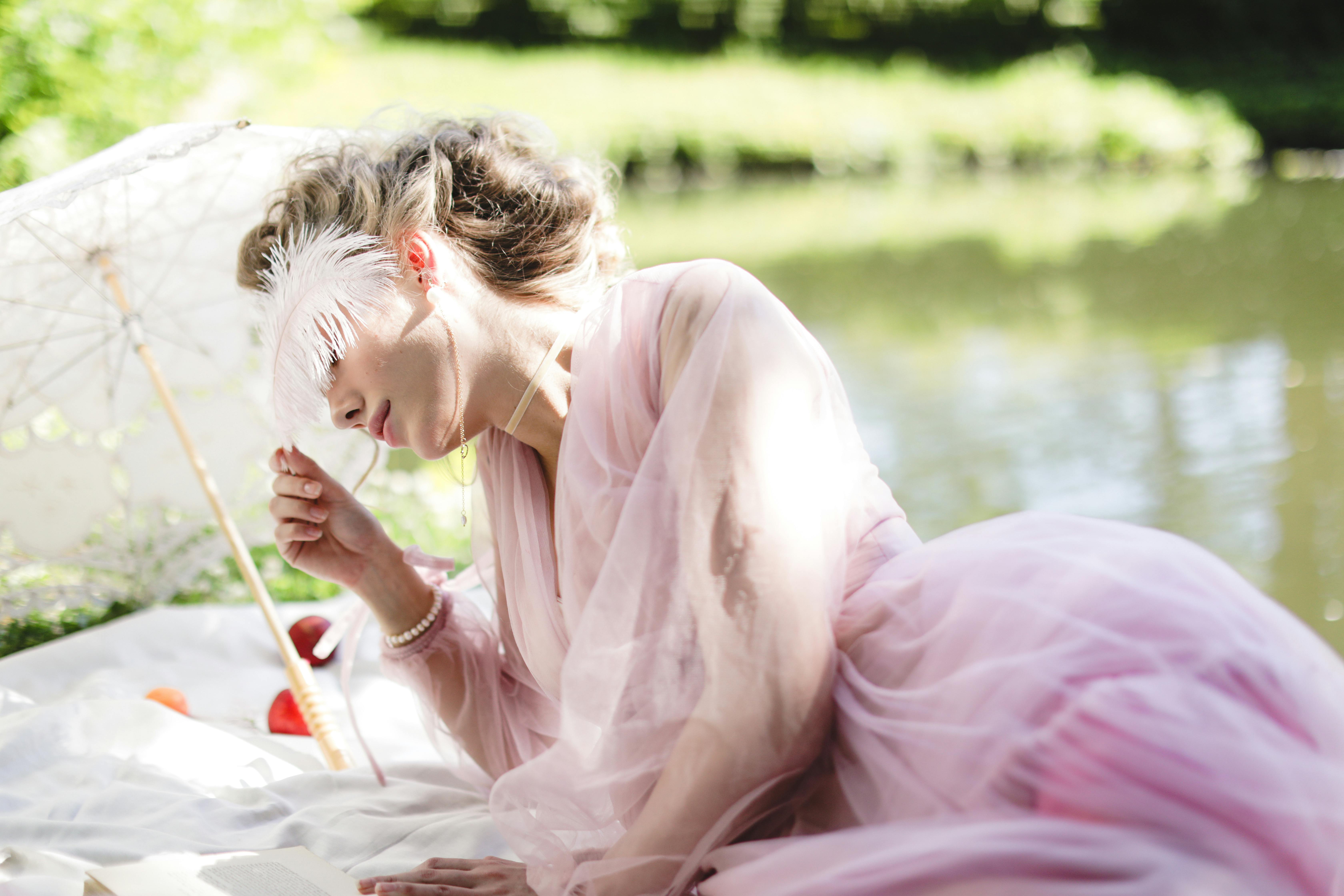 A person in a pink dress with a feather and umbrella by a lake.