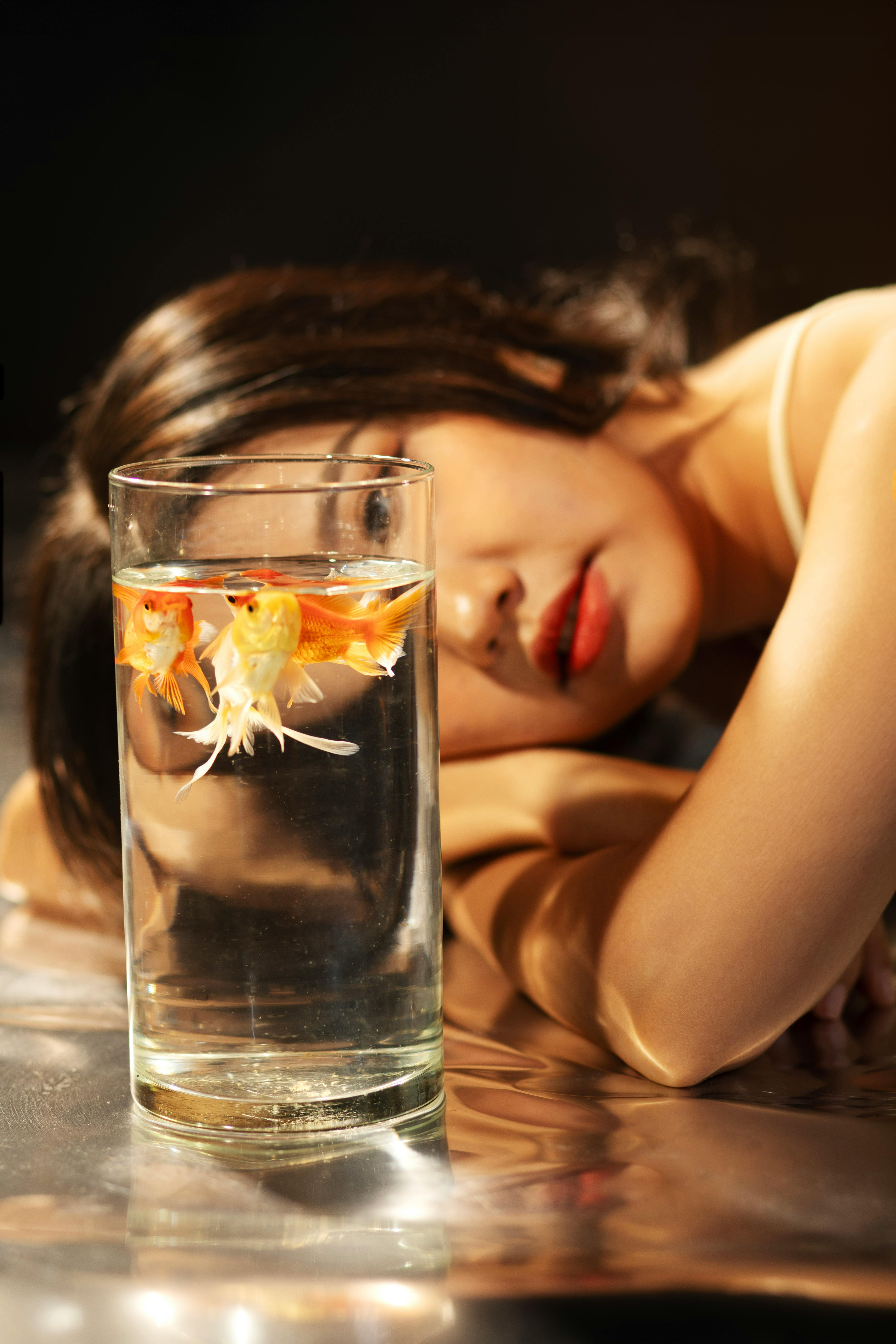 Person lying beside a glass of water with goldfish inside.