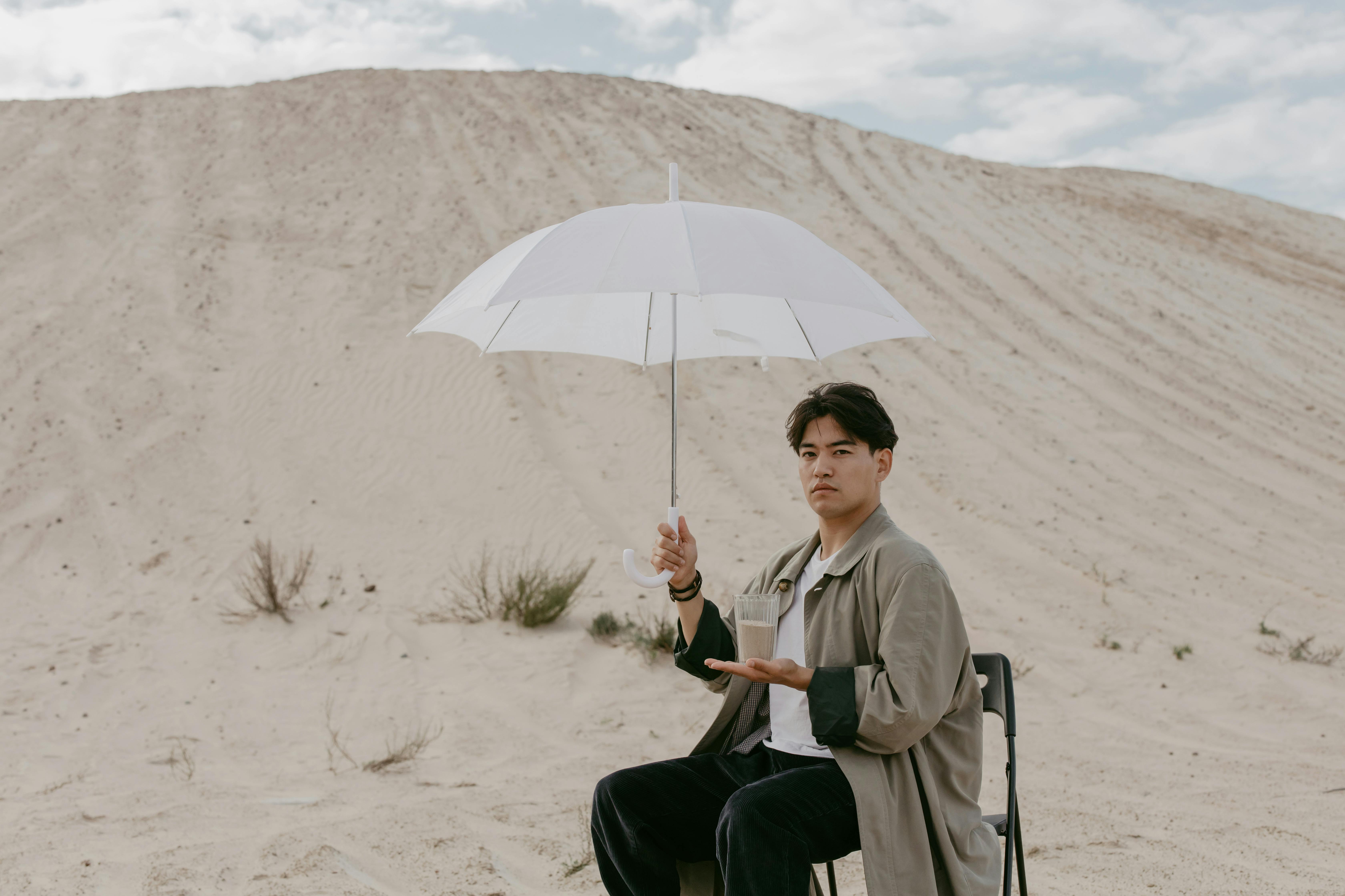 Person sitting in desert holding umbrella and glass of water.