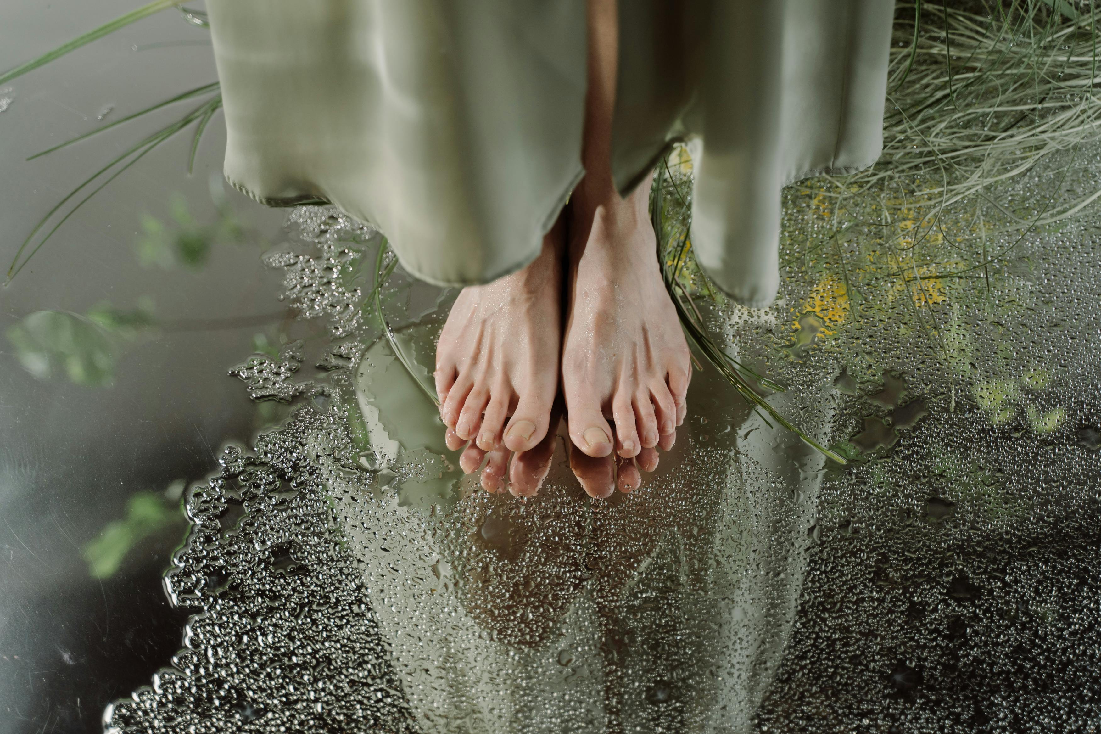 Bare feet on wet surface with reflection, surrounded by greenery.