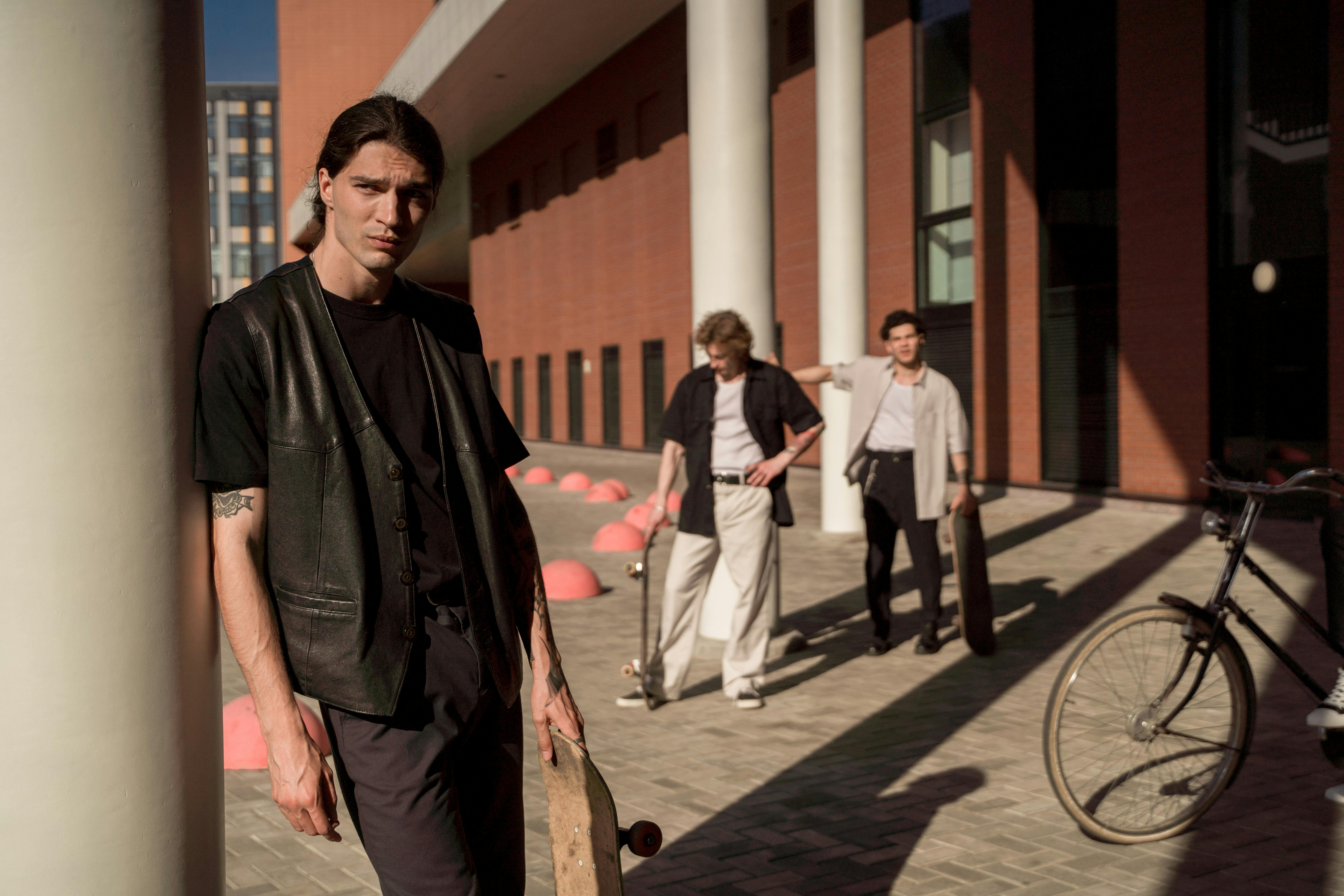 Three young men with skateboards and a bicycle outside.