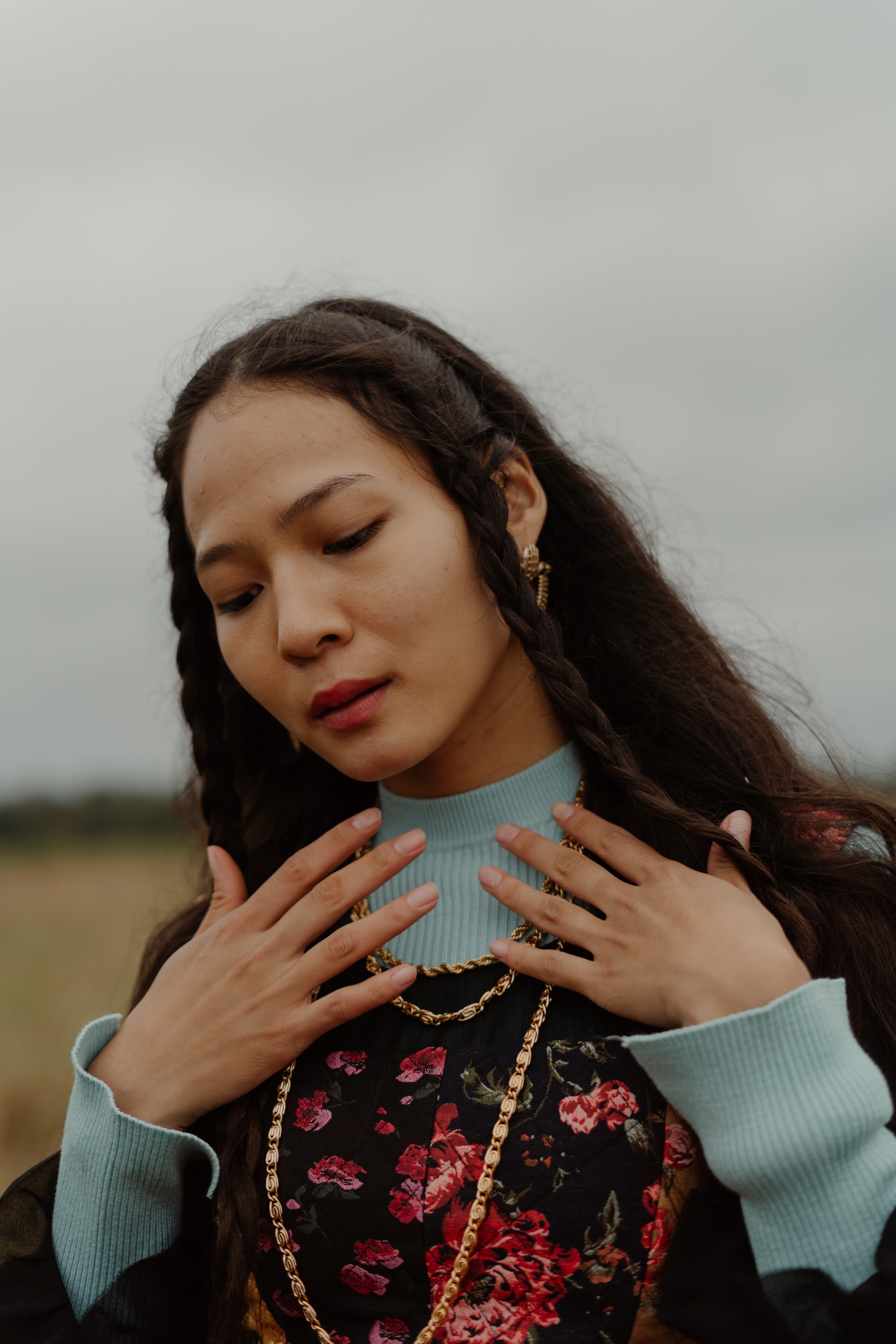 Person with braided hair, floral dress, gold chains, outdoors, thoughtful.