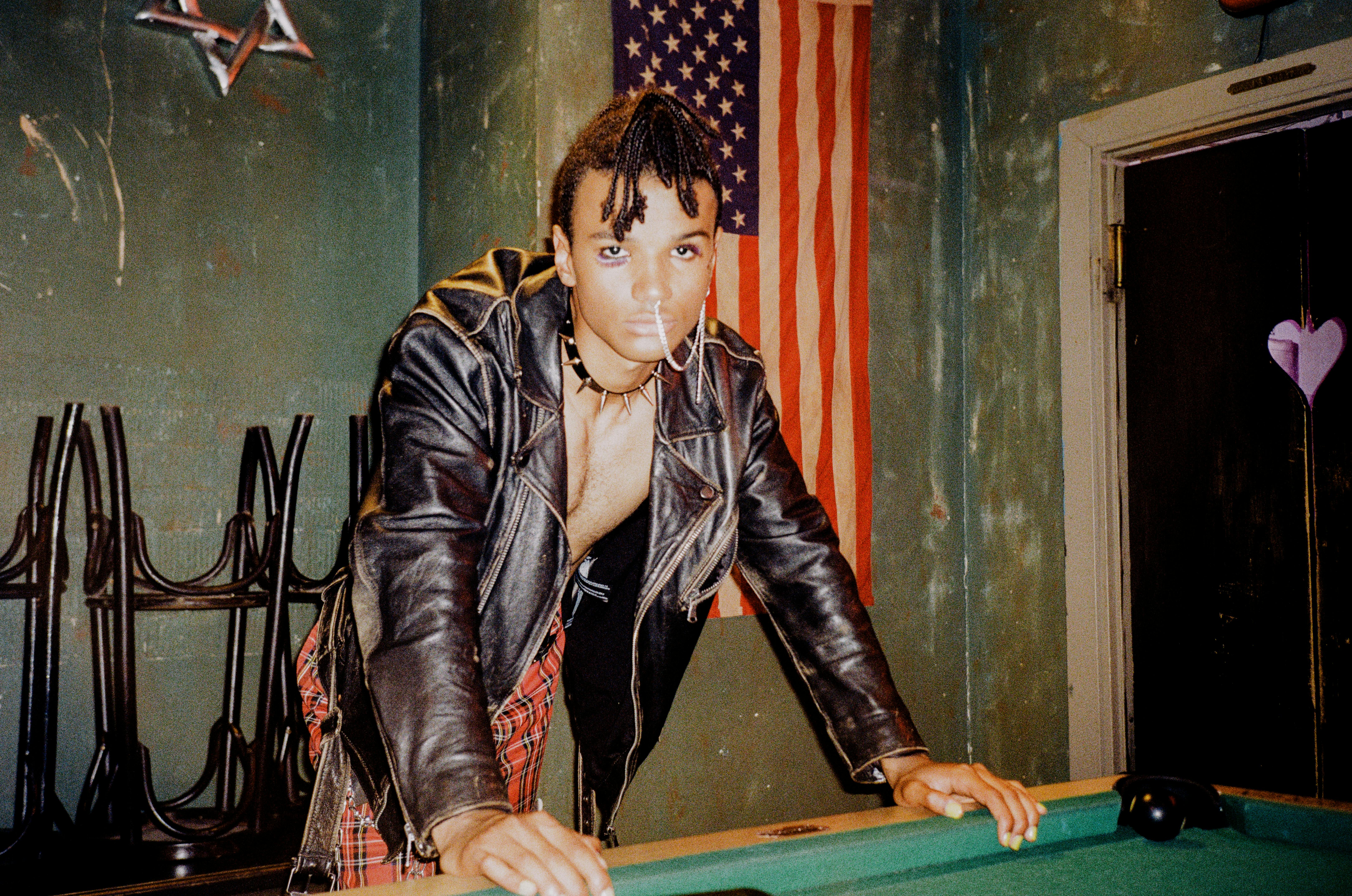 Person in leather jacket leans on pool table with American flag.