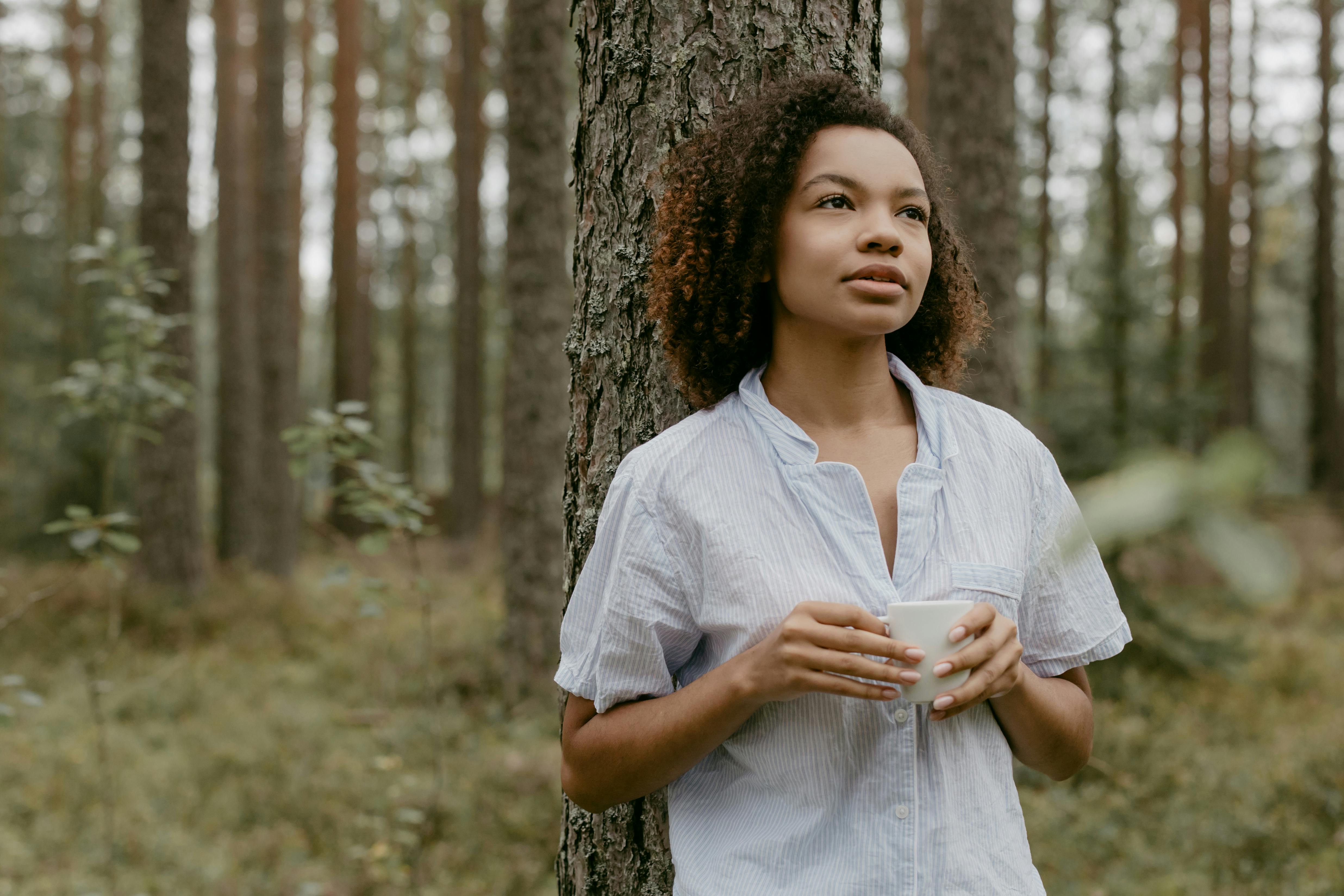 Person standing by tree in forest, holding a white mug.
