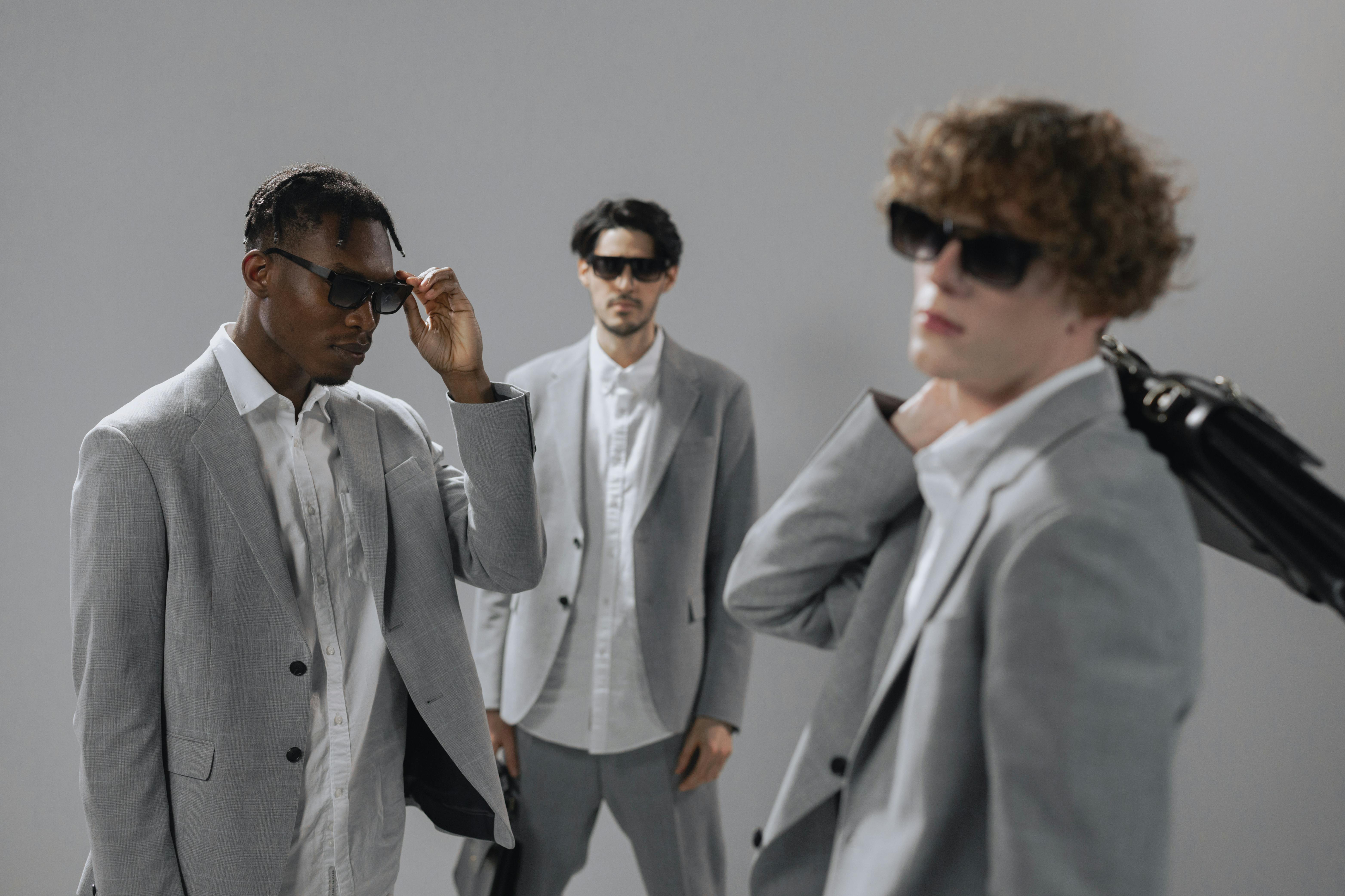 Three men in grey suits and sunglasses pose against a grey background.