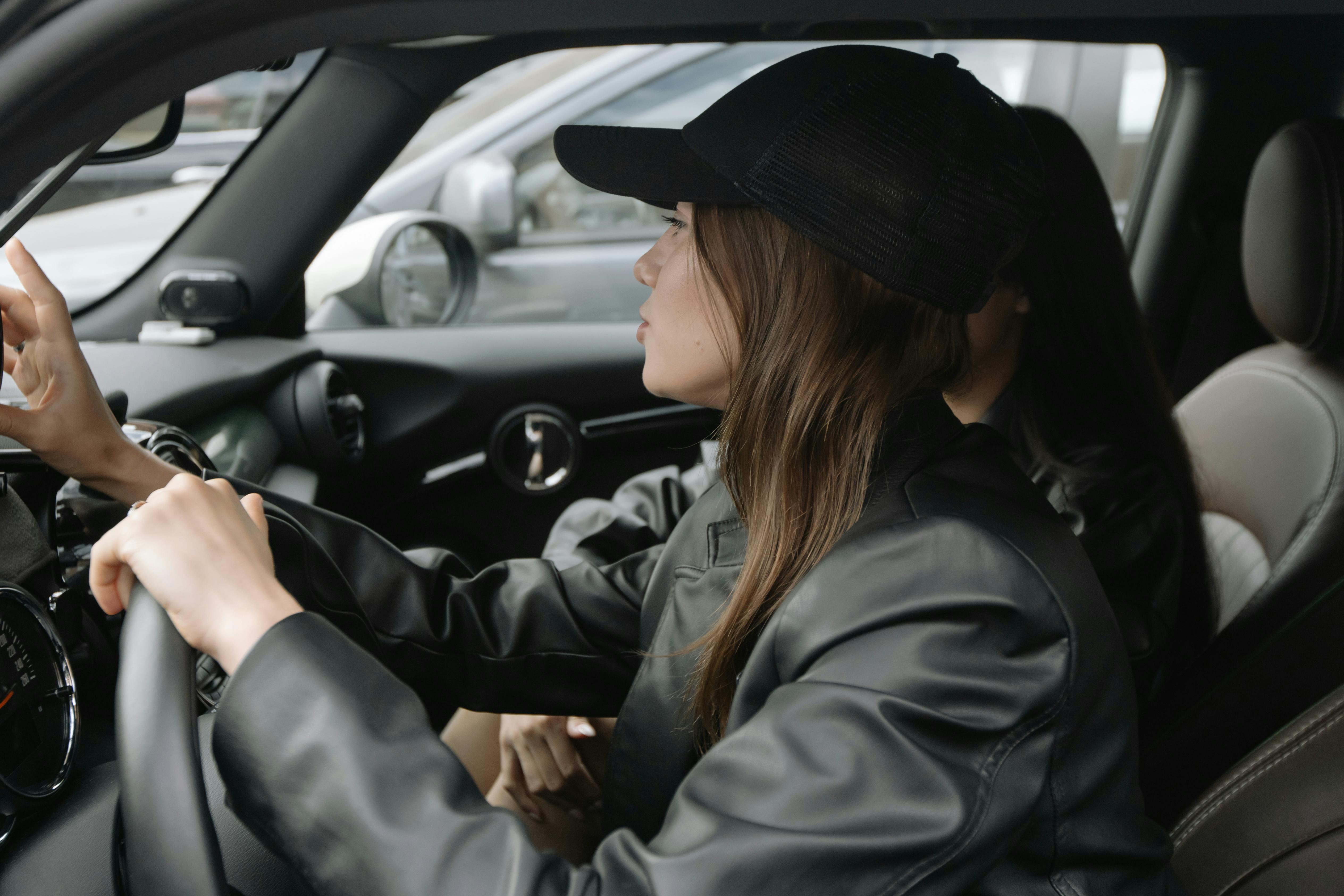 Person in black cap and jacket driving car, touching dashboard.