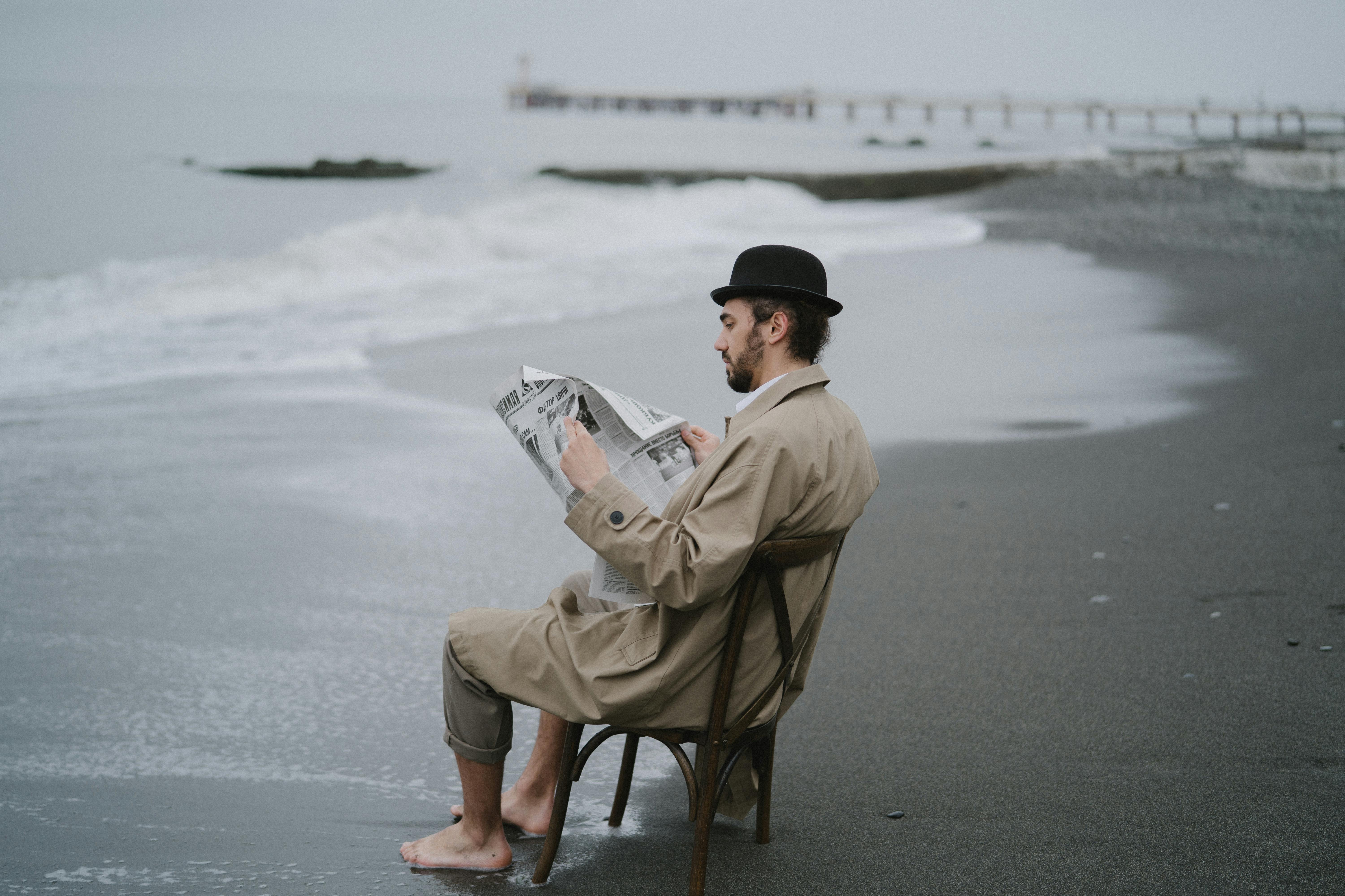 Person sitting on a chair on a beach reading a newspaper.