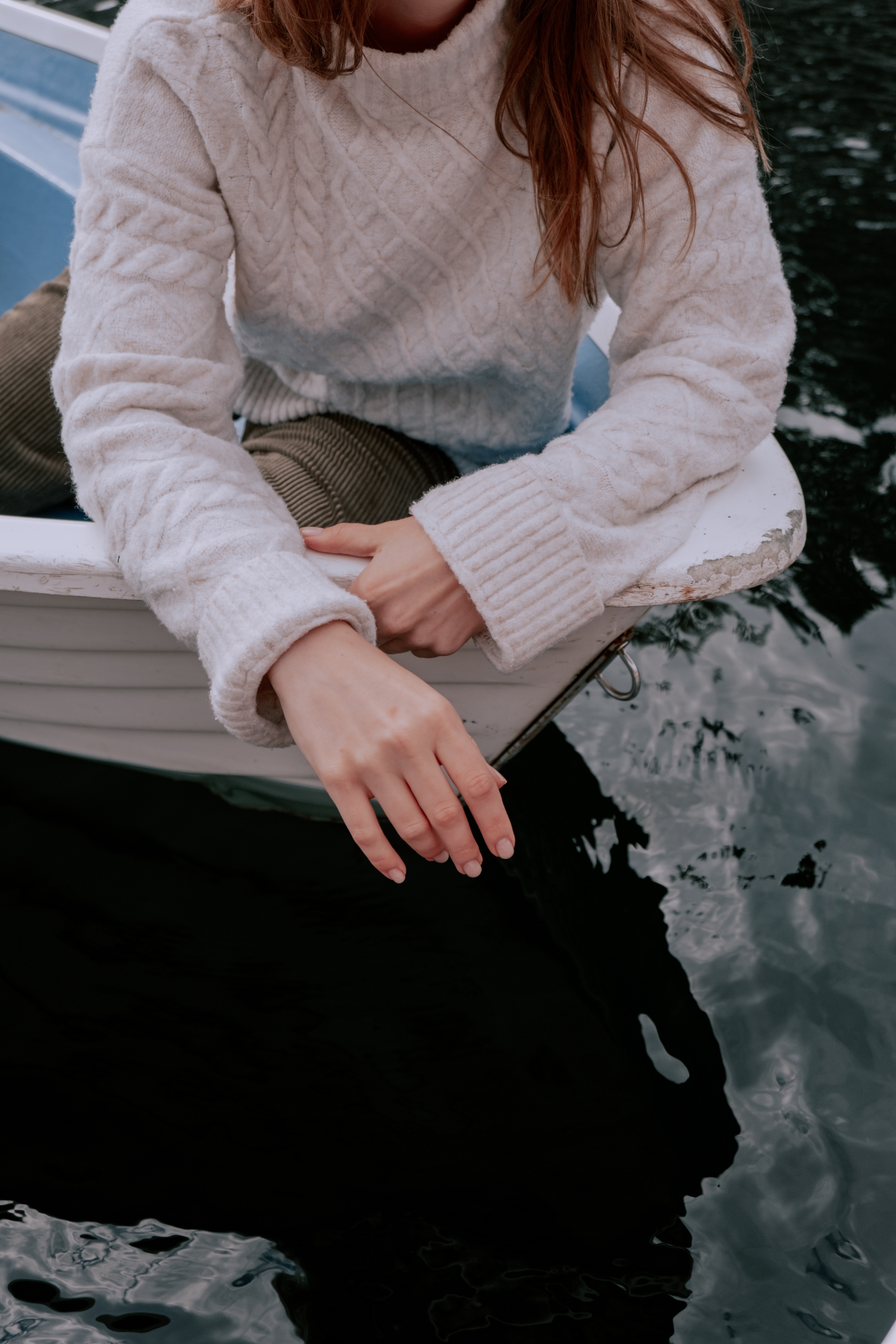 Person in cozy sweater sitting on boat, hand over water.