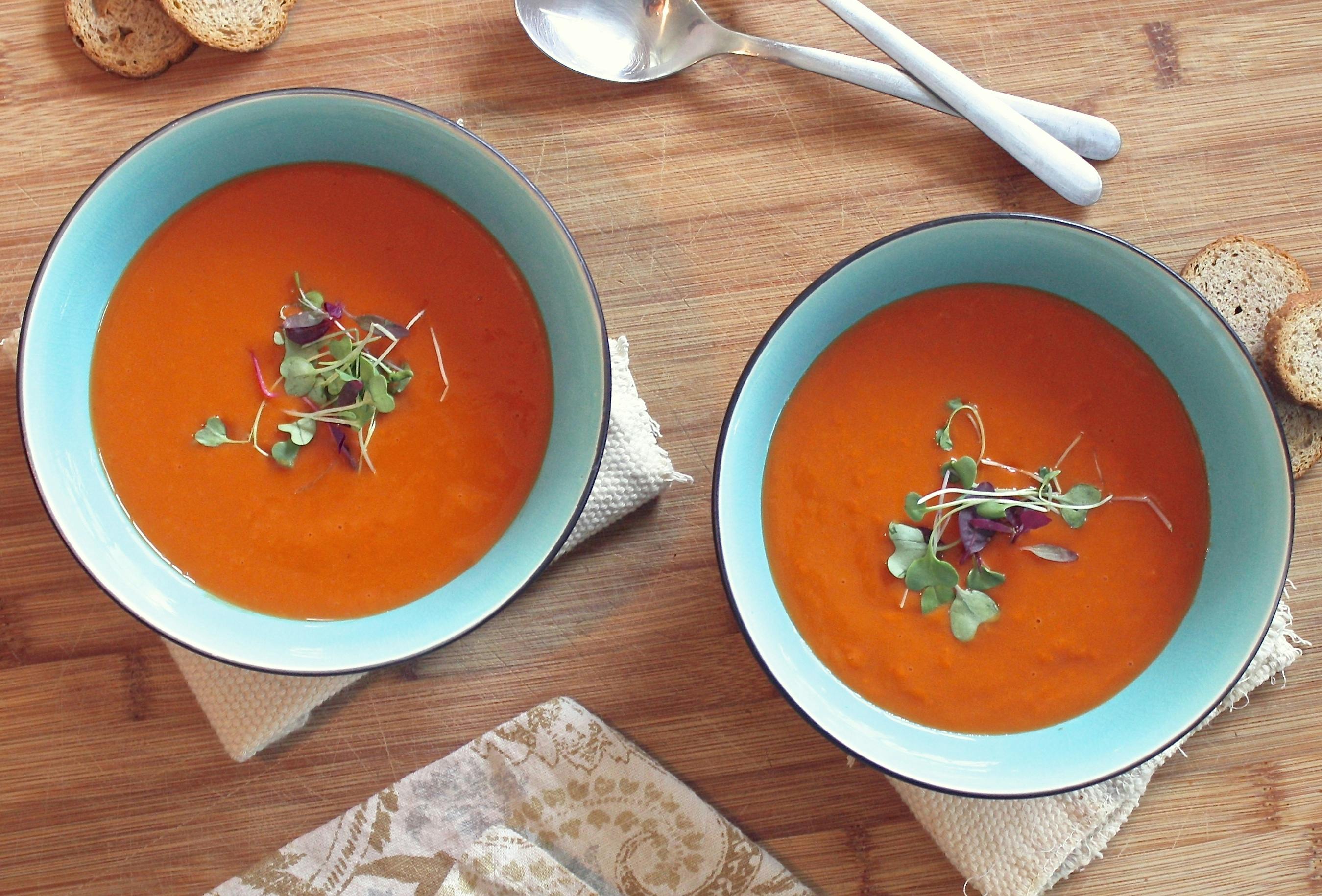 Two bowls of tomato soup with garnish, bread, and spoons.