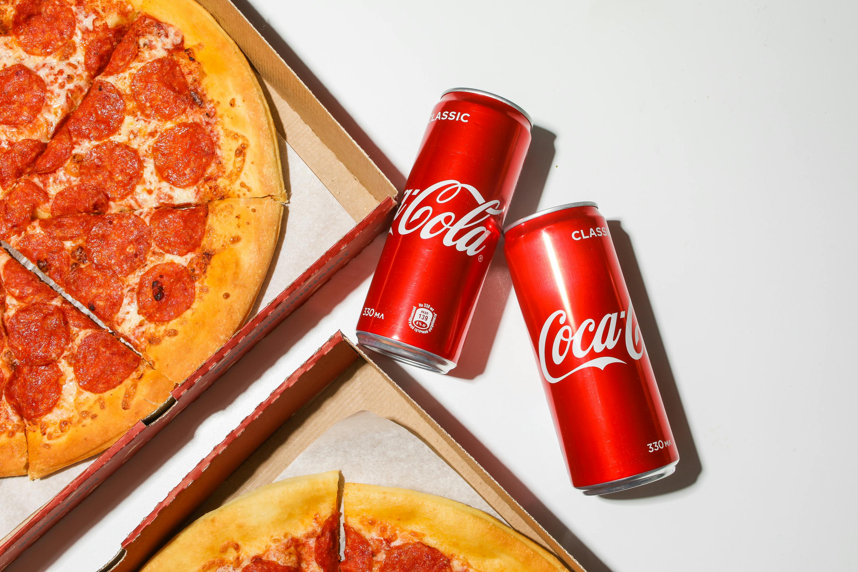 Two pepperoni pizzas in boxes with two cans of Coca-Cola.