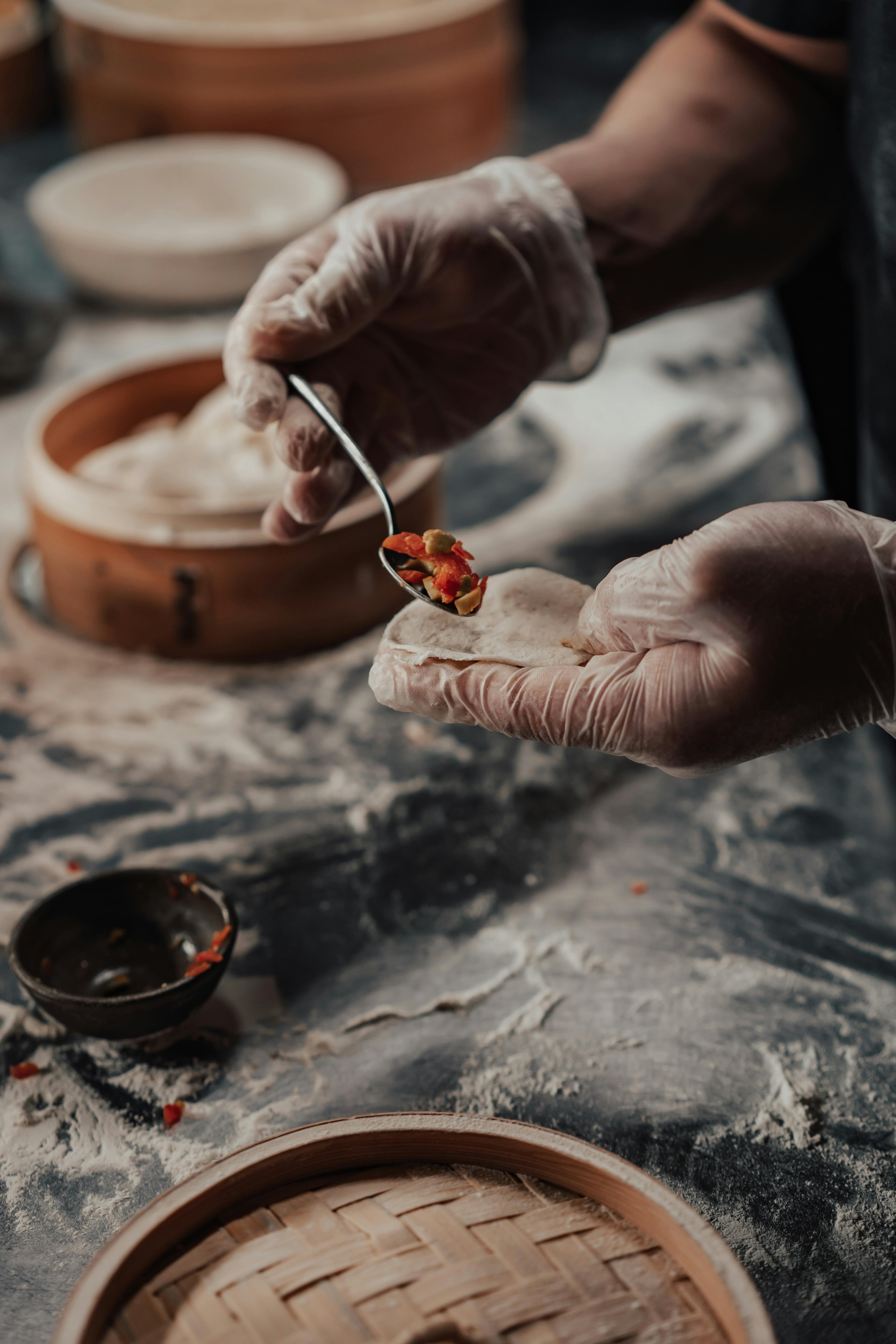 Gloved hands preparing dumplings with filling on a floured surface.