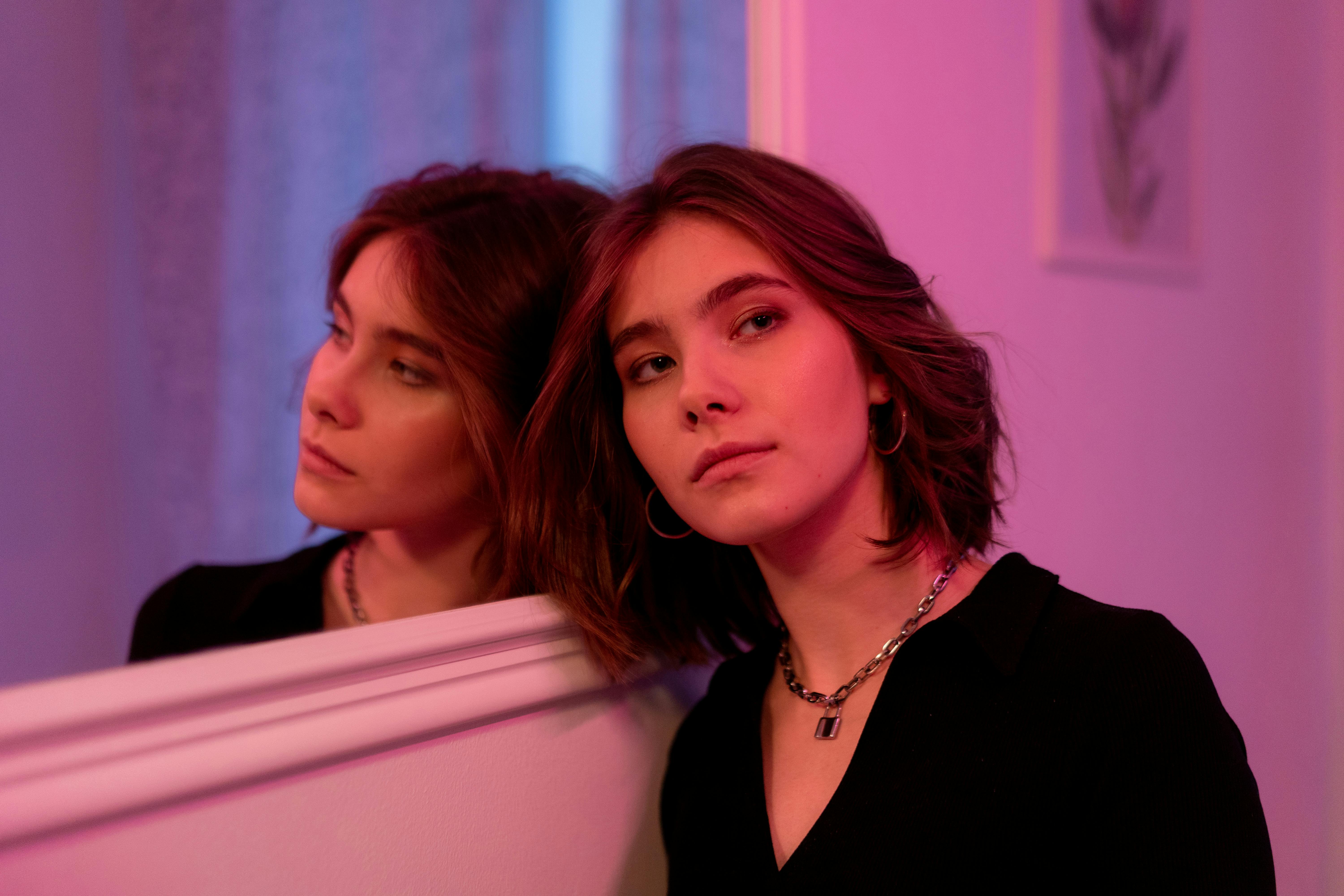 Person leaning against mirror, pink lighting, thoughtful expression, casual outfit.