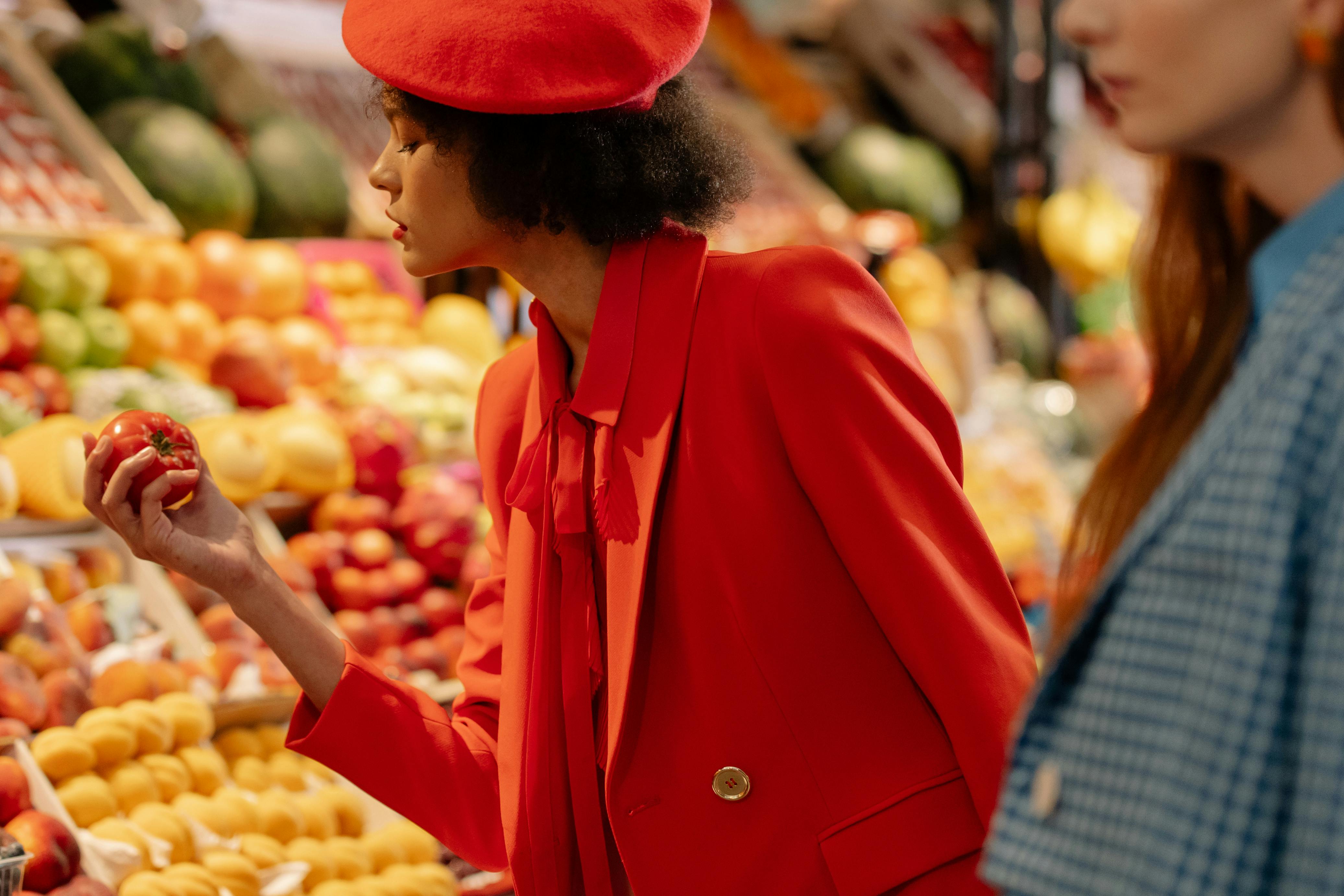 Person in red outfit at a fruit stand holding a tomato.