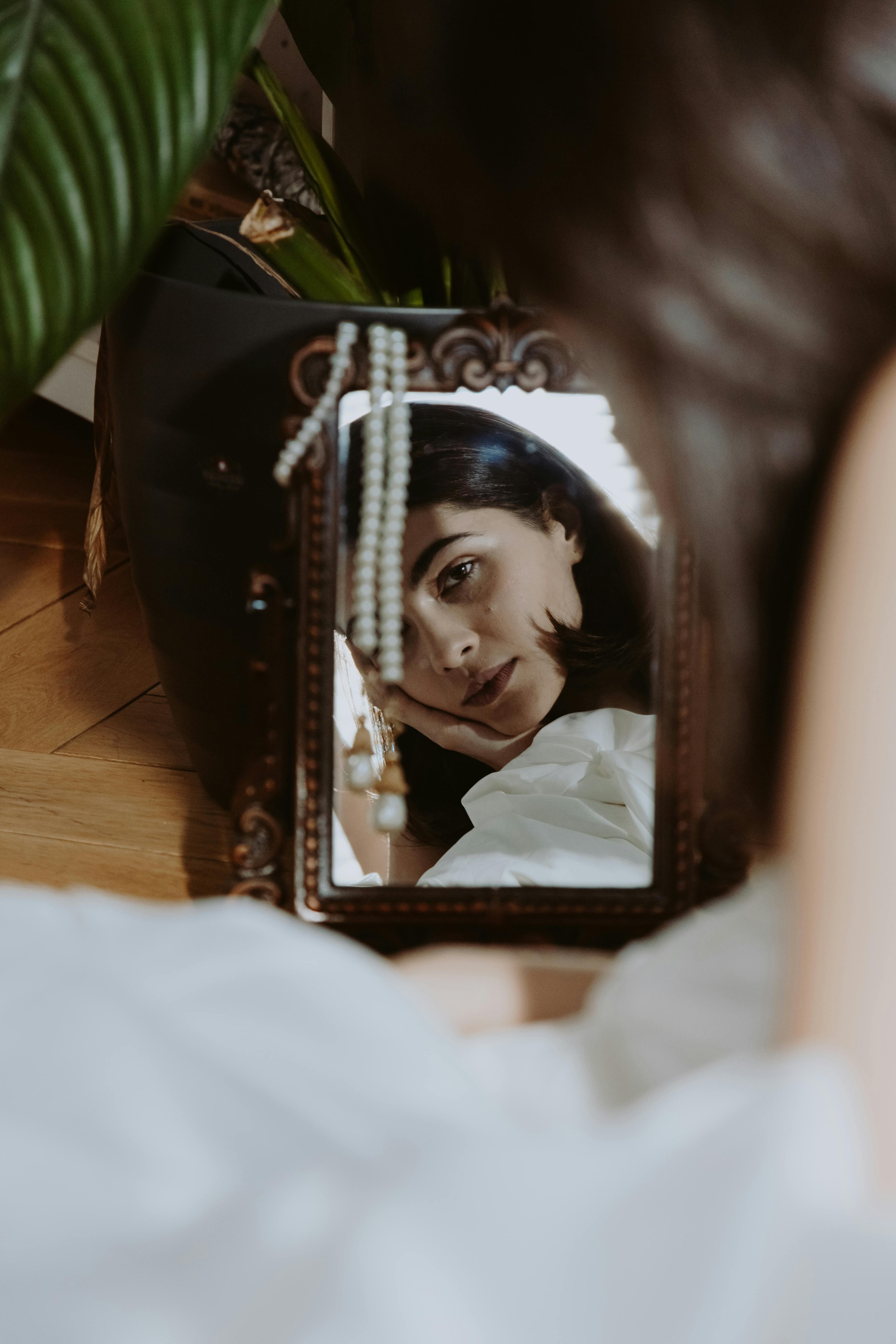 Person's reflection in ornate mirror, pearls hanging, indoor setting.