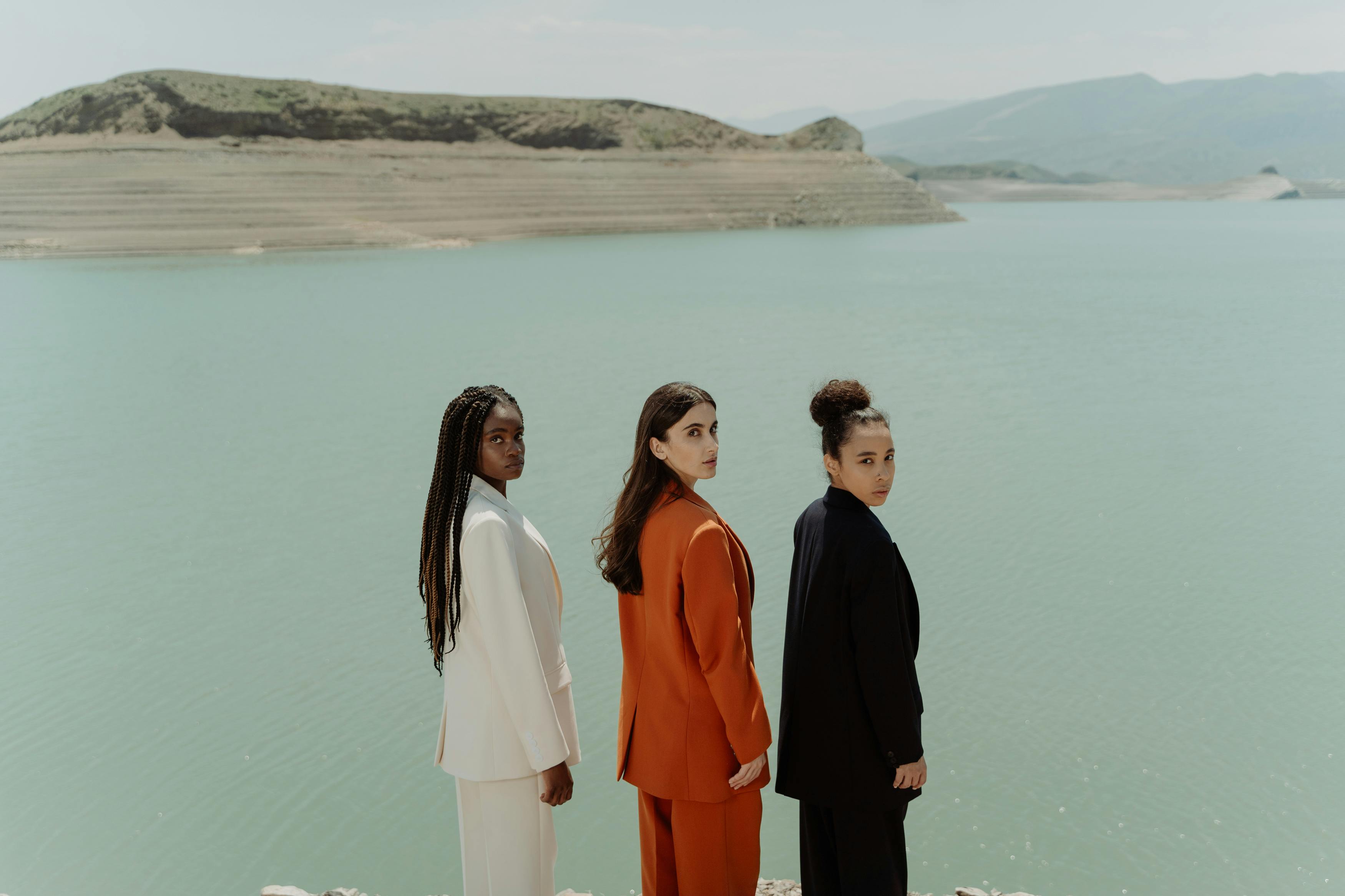 Three people in suits stand by a calm lake with hills.