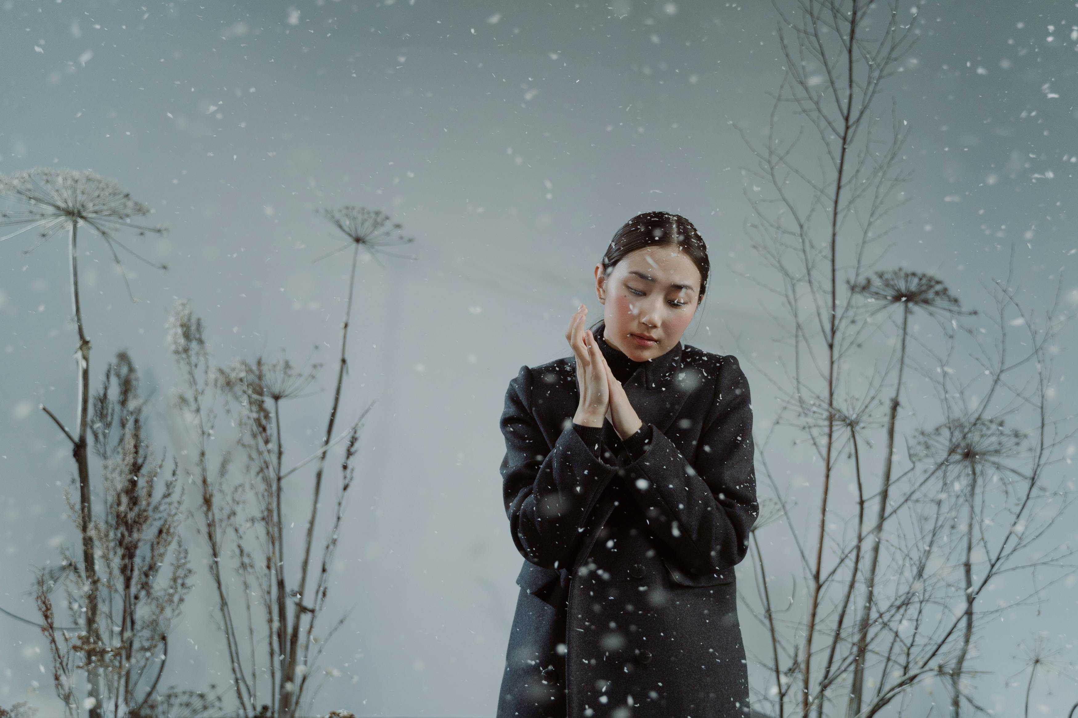 Person standing in falling snow, surrounded by bare winter plants.