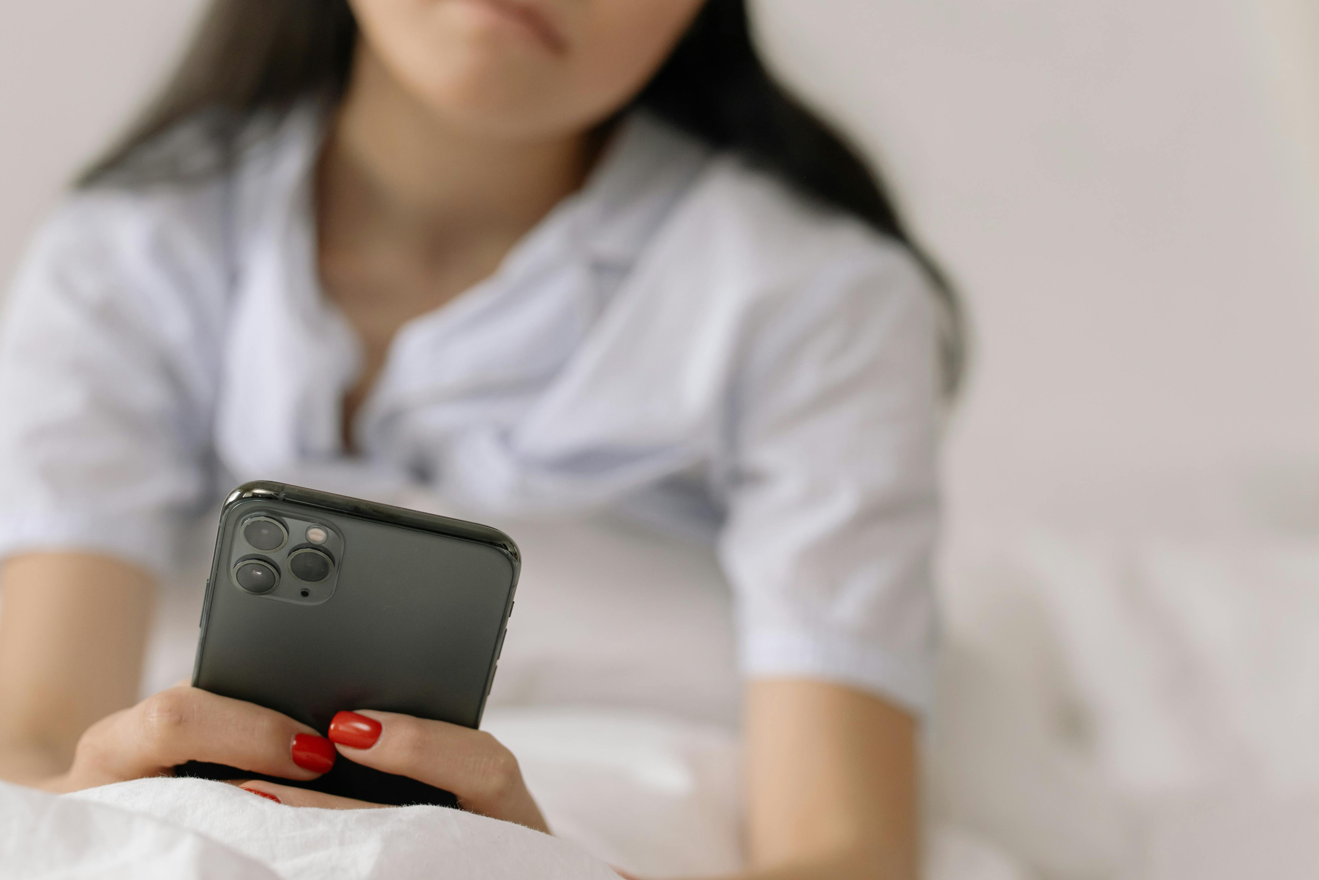 Person lying on bed, holding smartphone with red painted nails.