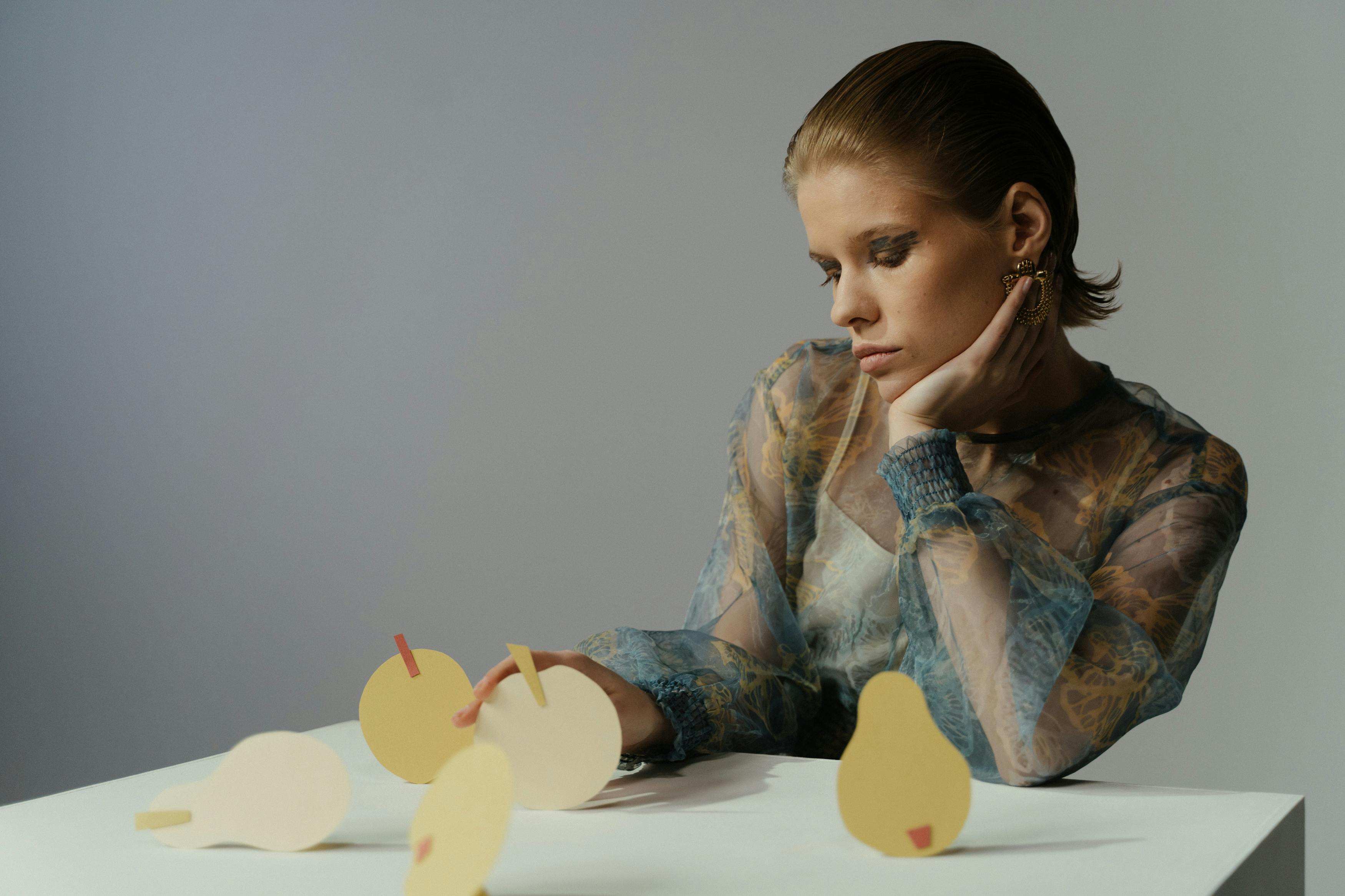 Person with hand on chin near paper fruit shapes on table.