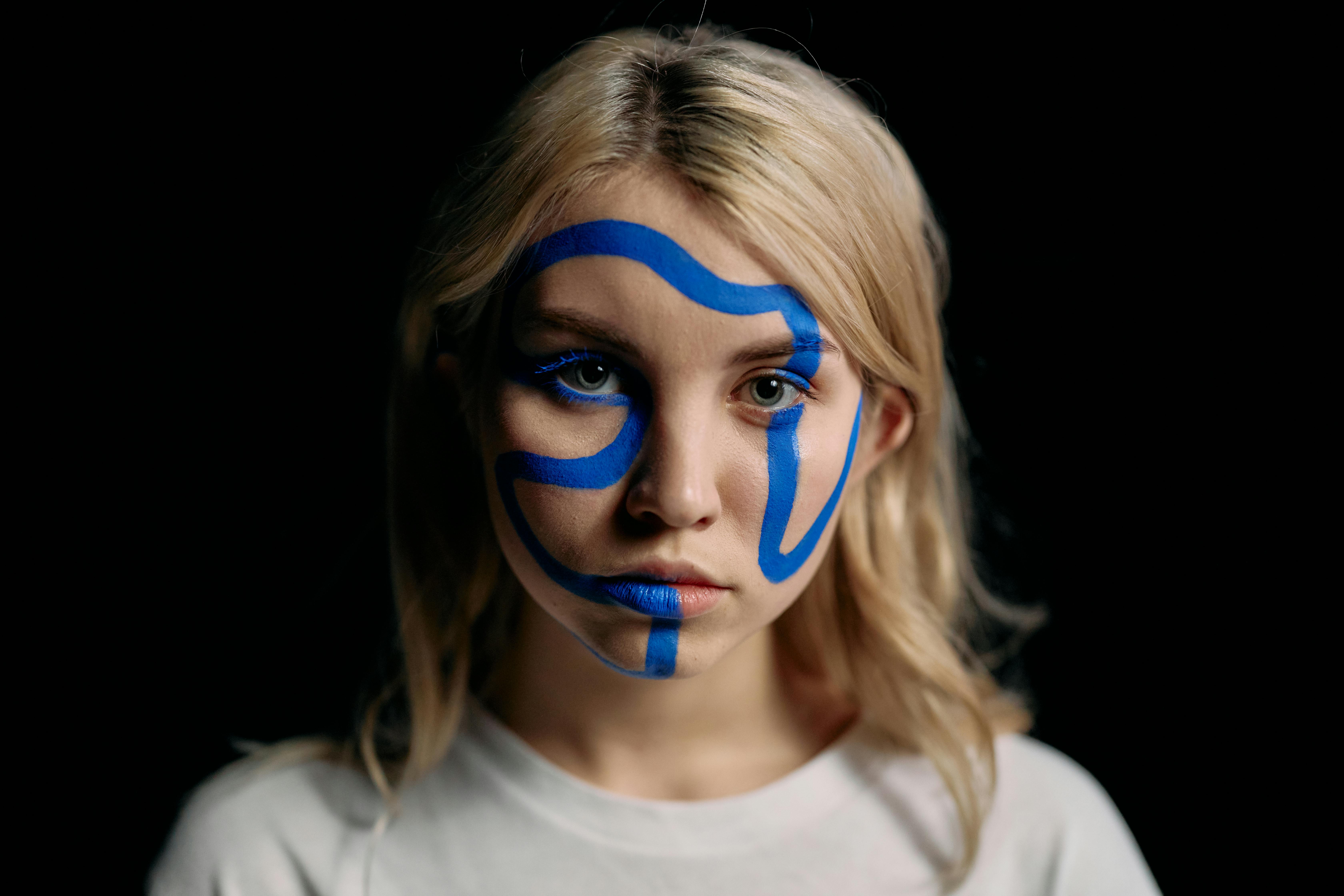 Person with abstract blue face paint, neutral expression, dark background.