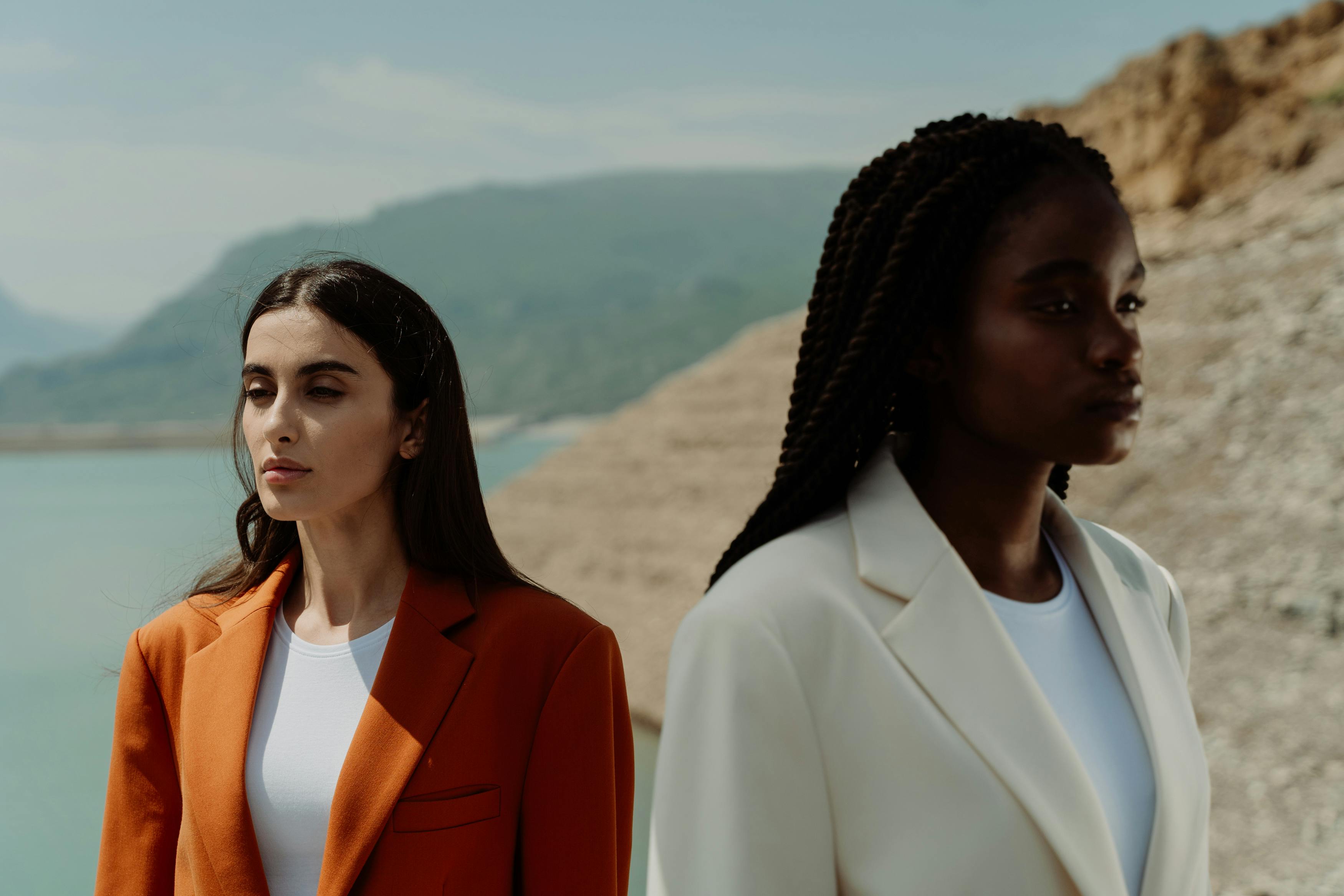 Two people in blazers stand against a backdrop of water and hills.