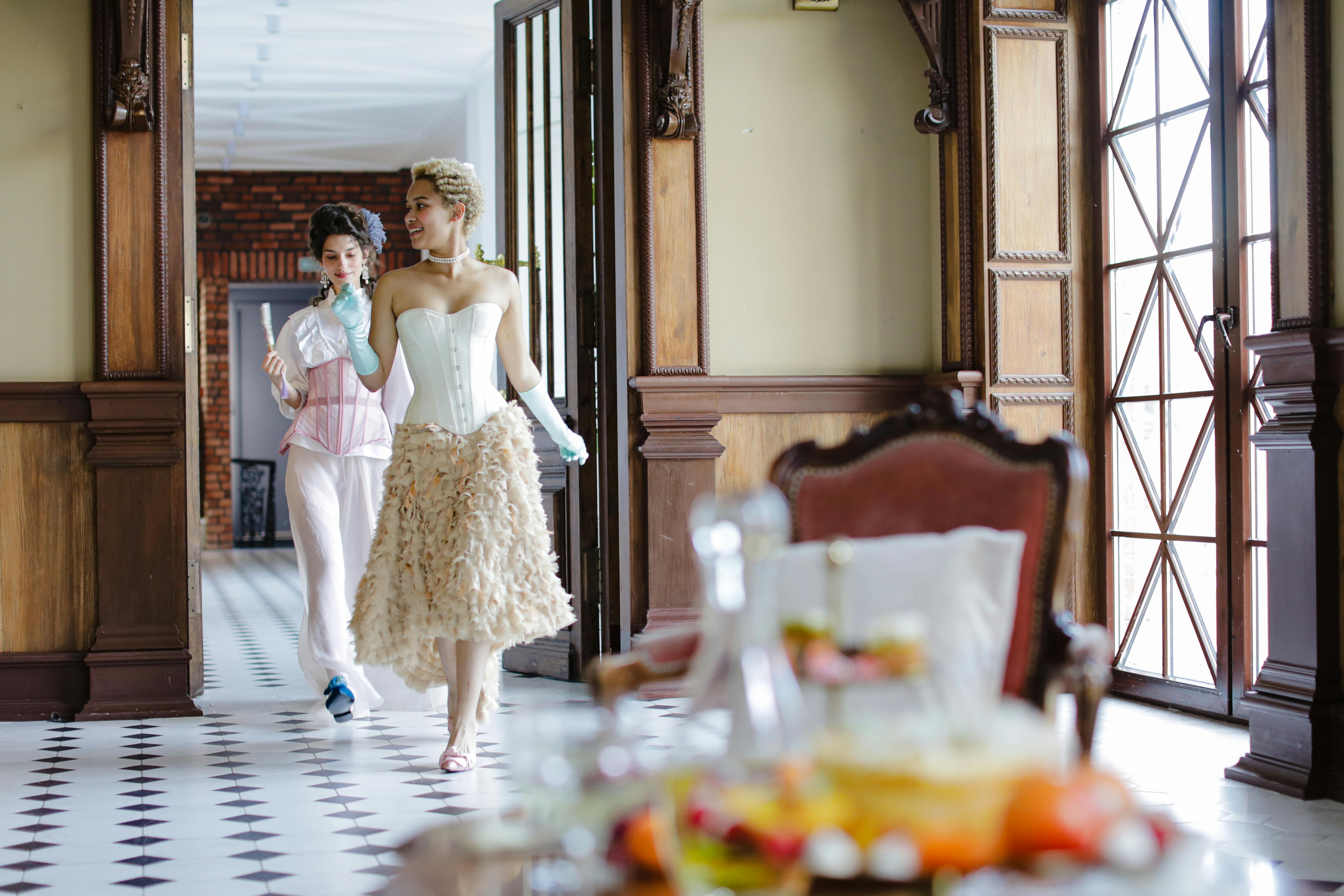 Two women in vintage attire walk through a grand hallway.