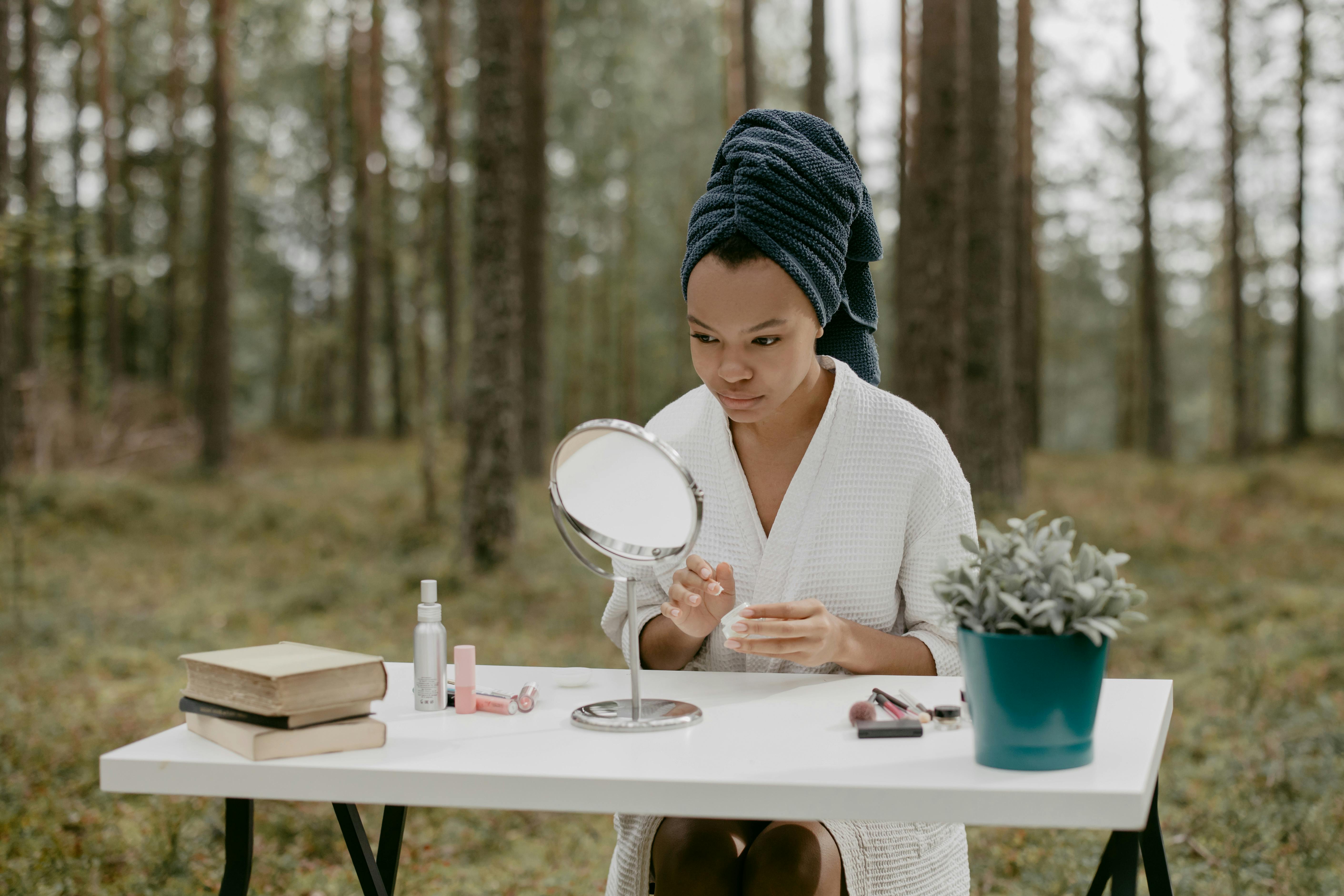 Person sitting at table in forest, applying skincare, mirror nearby.