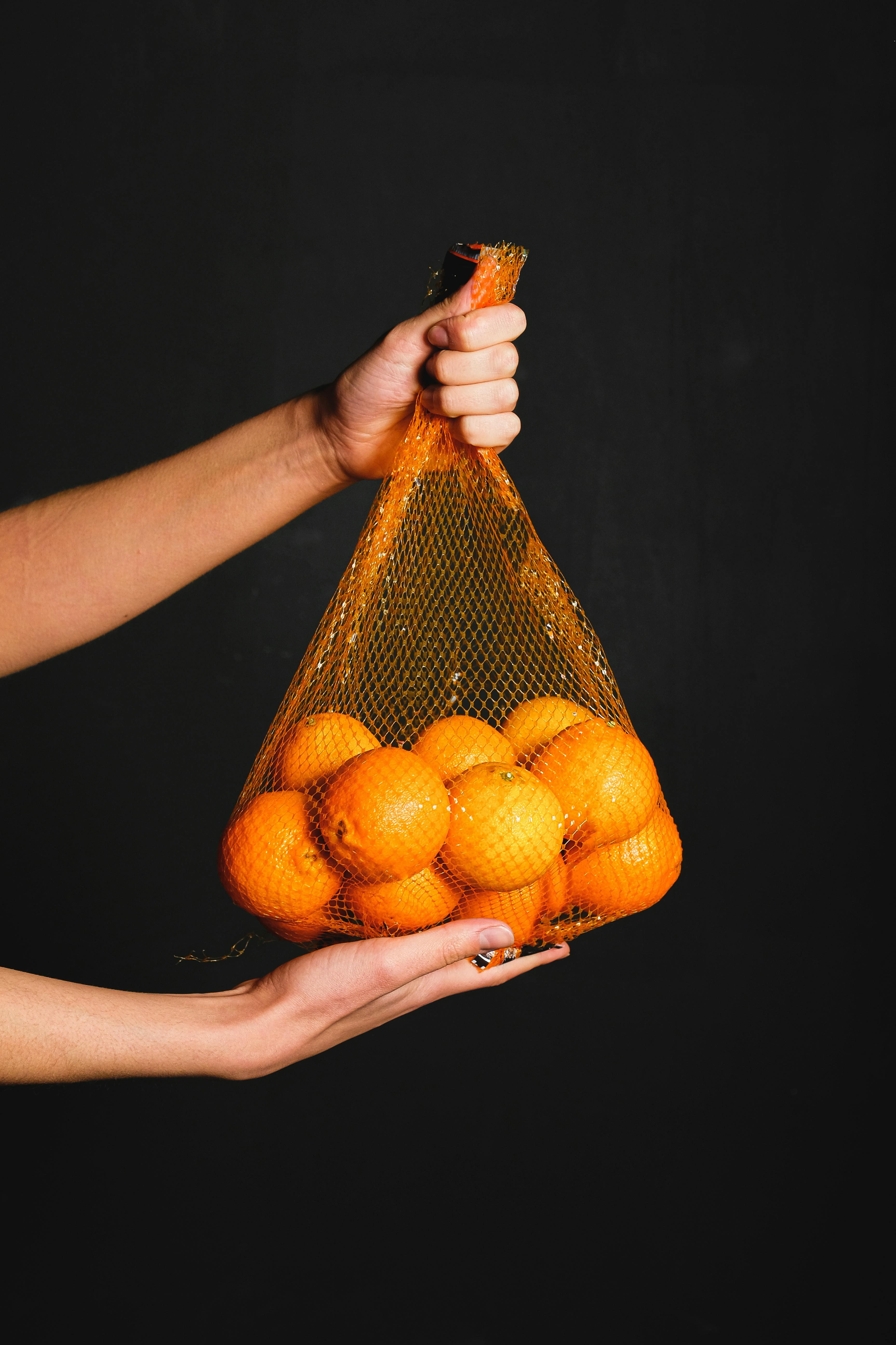 A hand holds a mesh bag filled with oranges, black background.