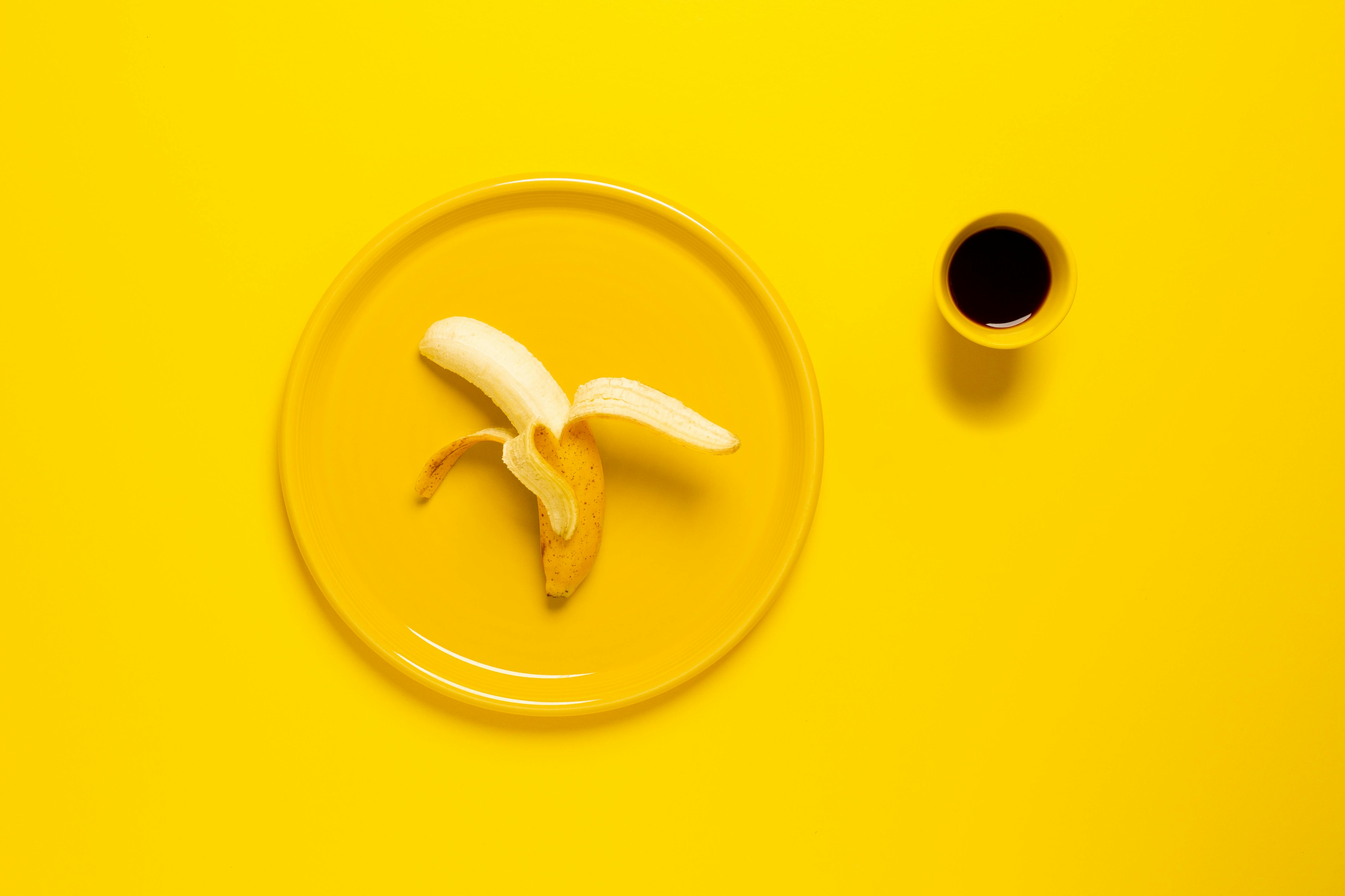 Peeled banana on yellow plate, cup of coffee, yellow background.