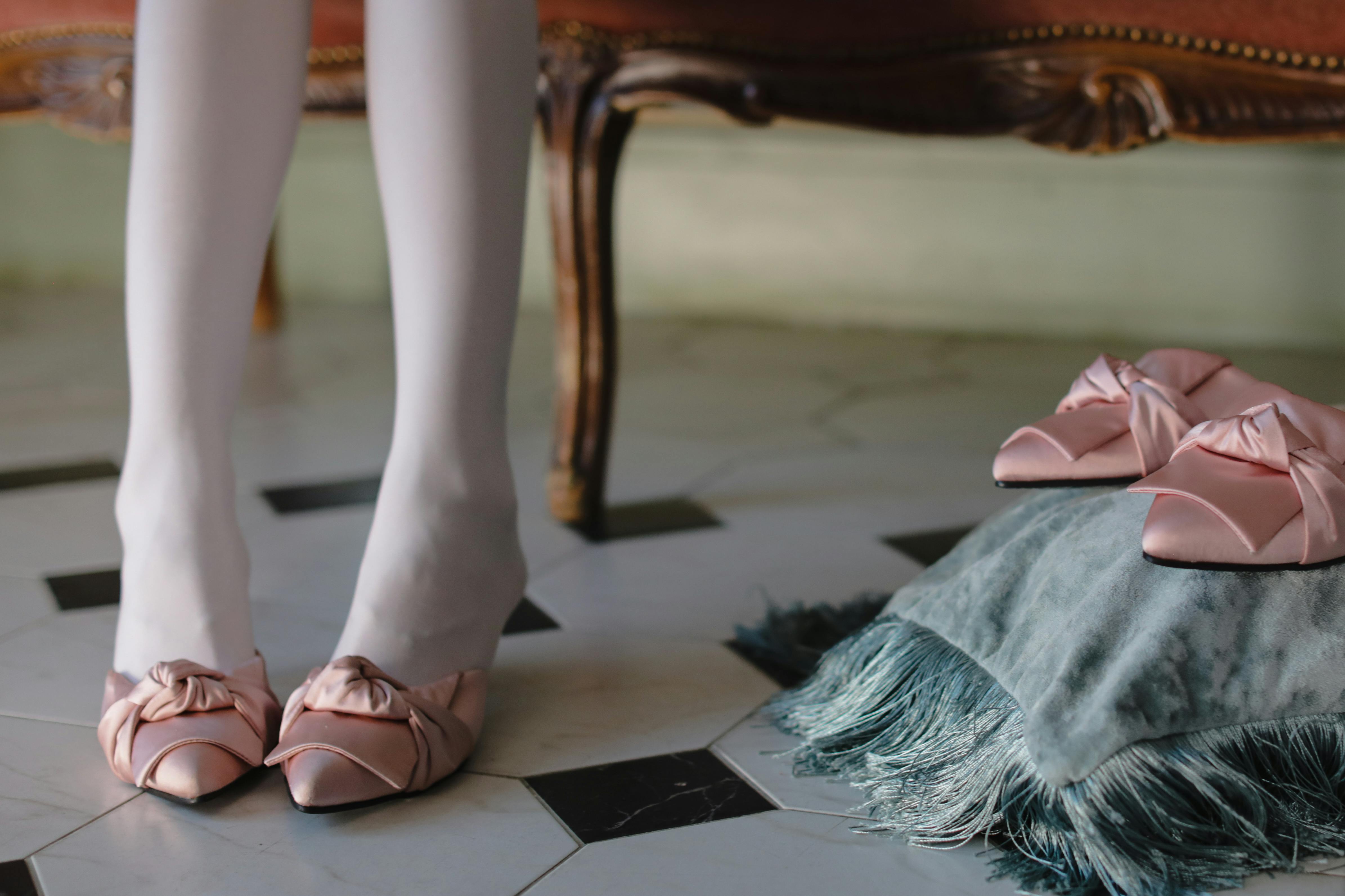 A person wearing white stockings and pink bow shoes on a tiled floor.