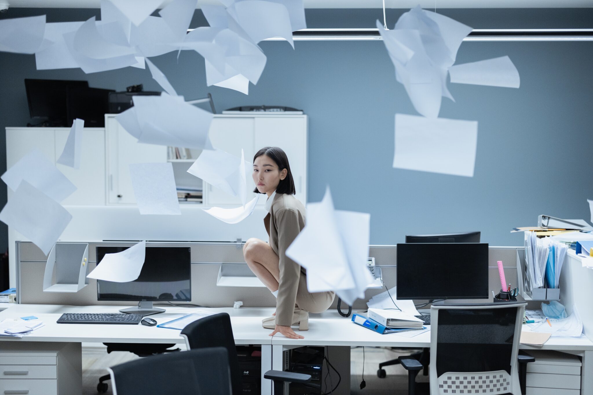 Person crouching on office desk, papers flying everywhere, chaotic scene.