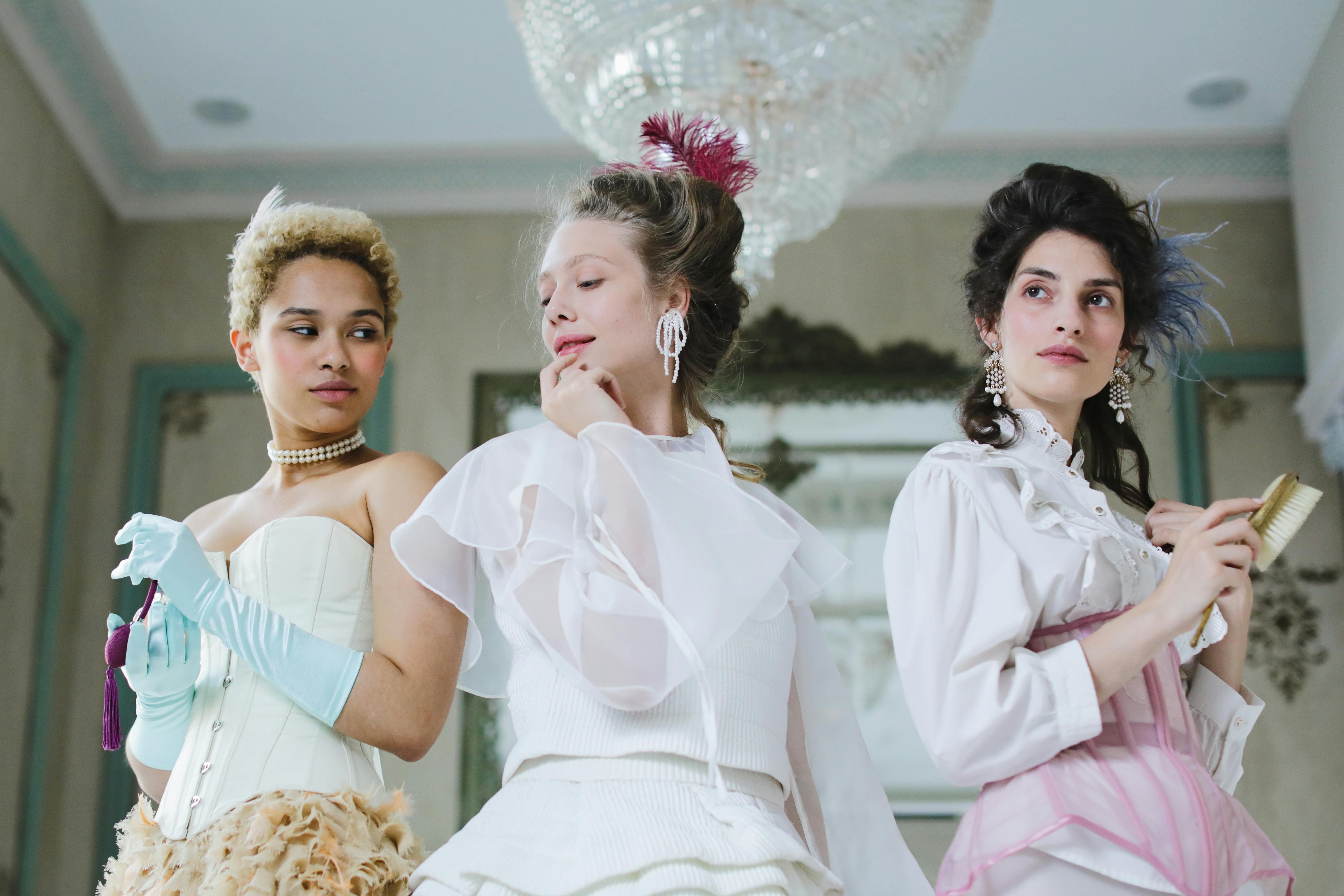 Three women in elaborate historical gowns stand in a luxurious room.
