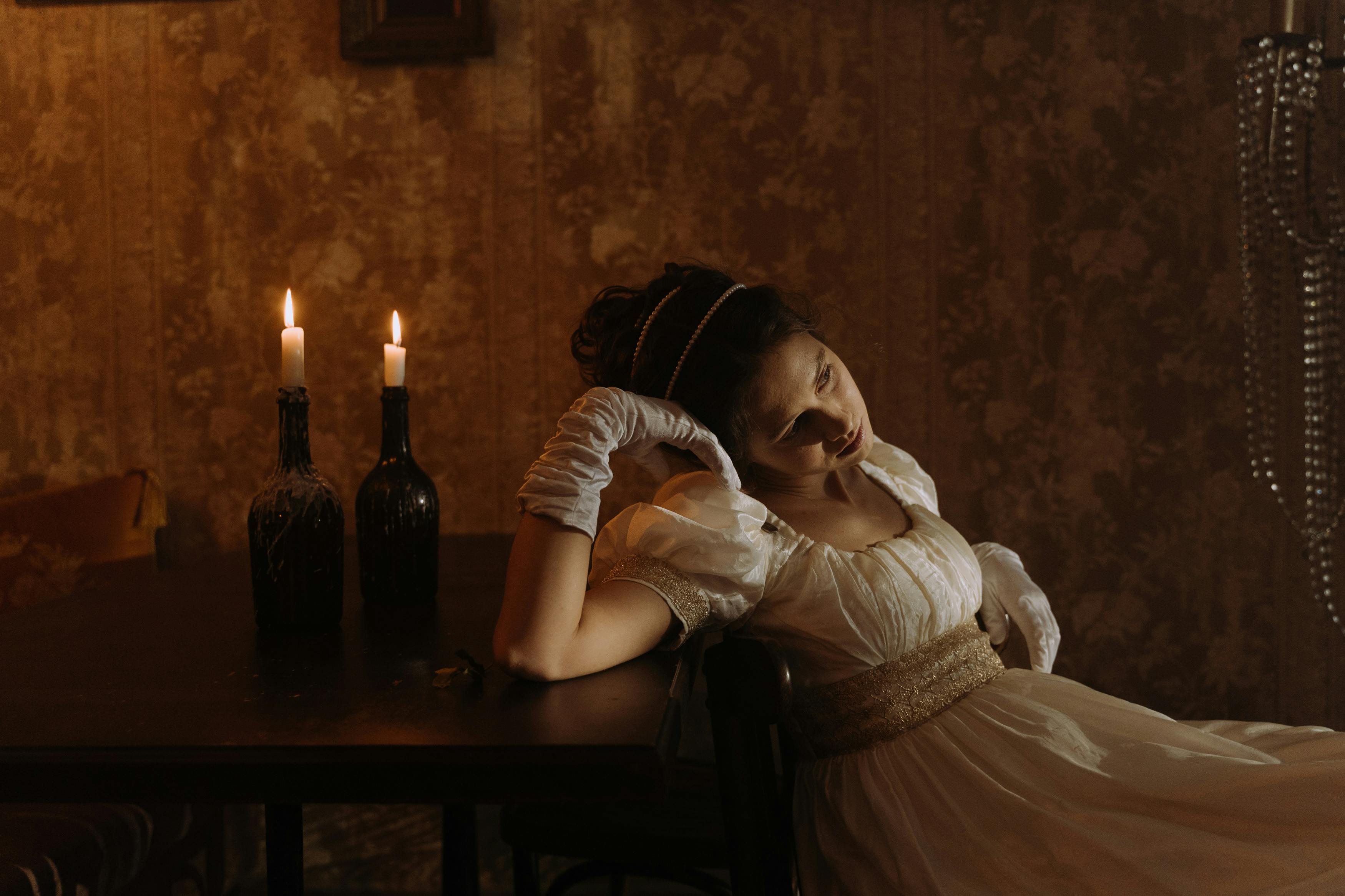 Person in vintage dress sits by candlelit table, looking contemplative.