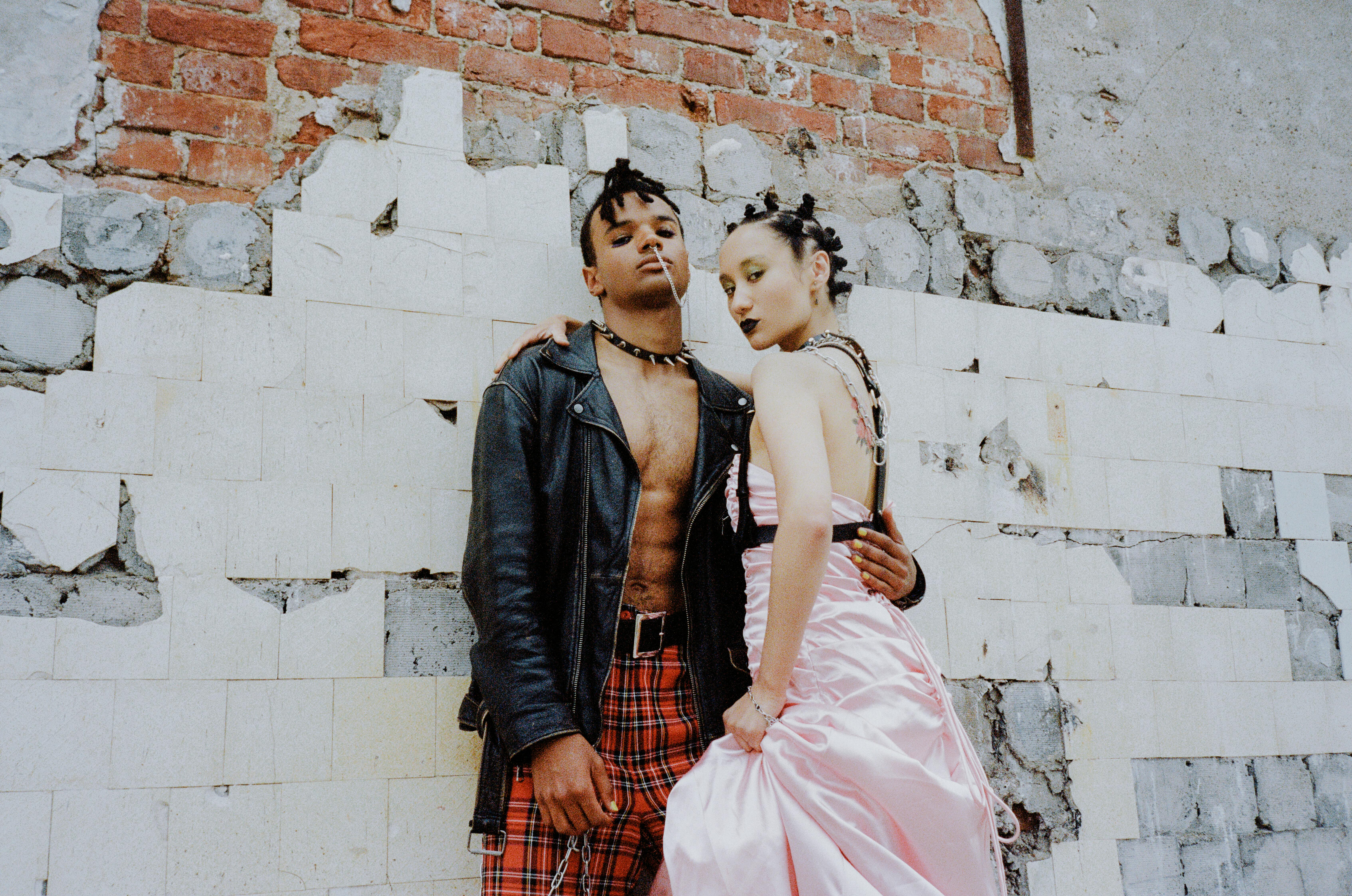 Two people in edgy fashion stand against a distressed brick wall.