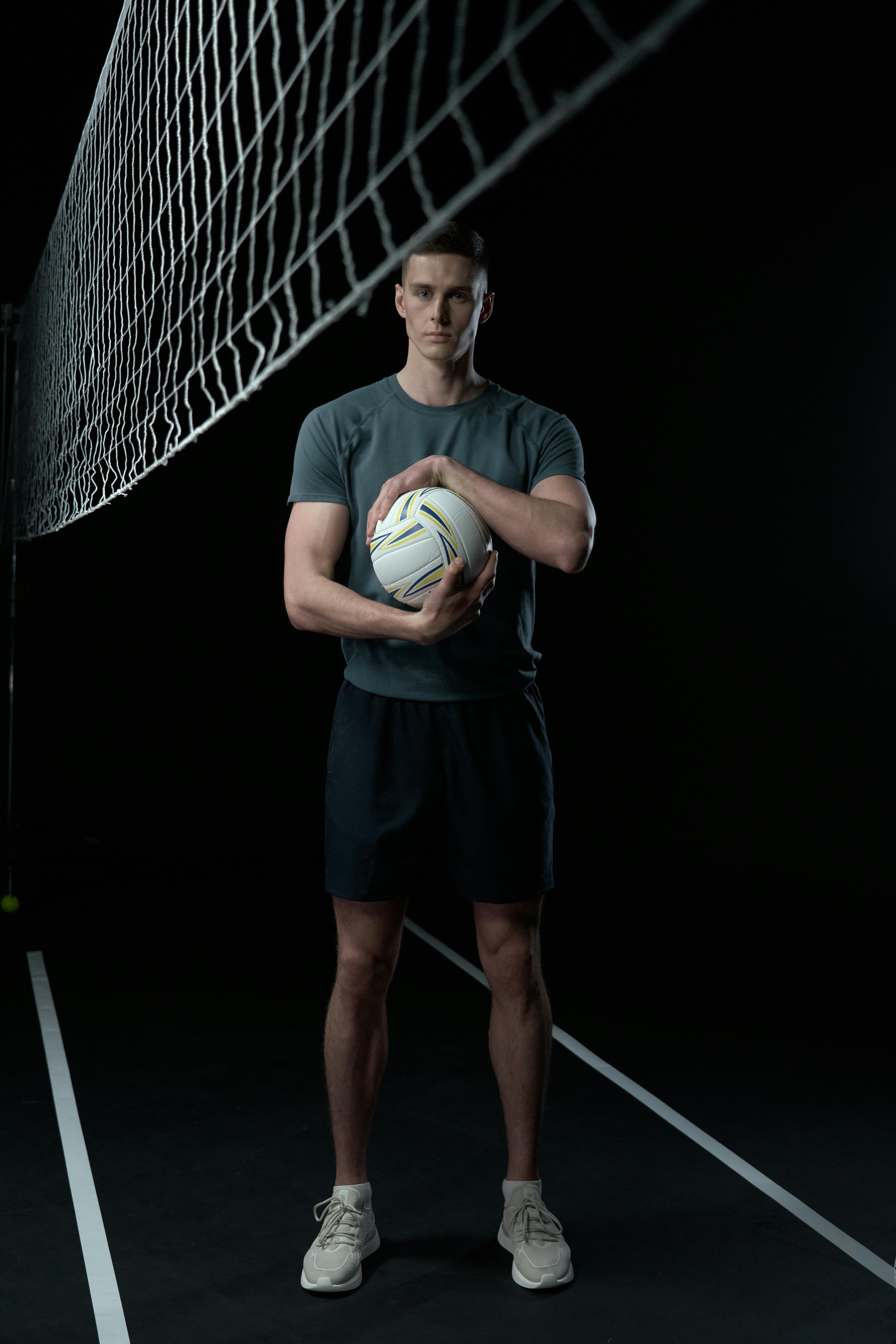 Person standing on volleyball court holding a volleyball, dark background.