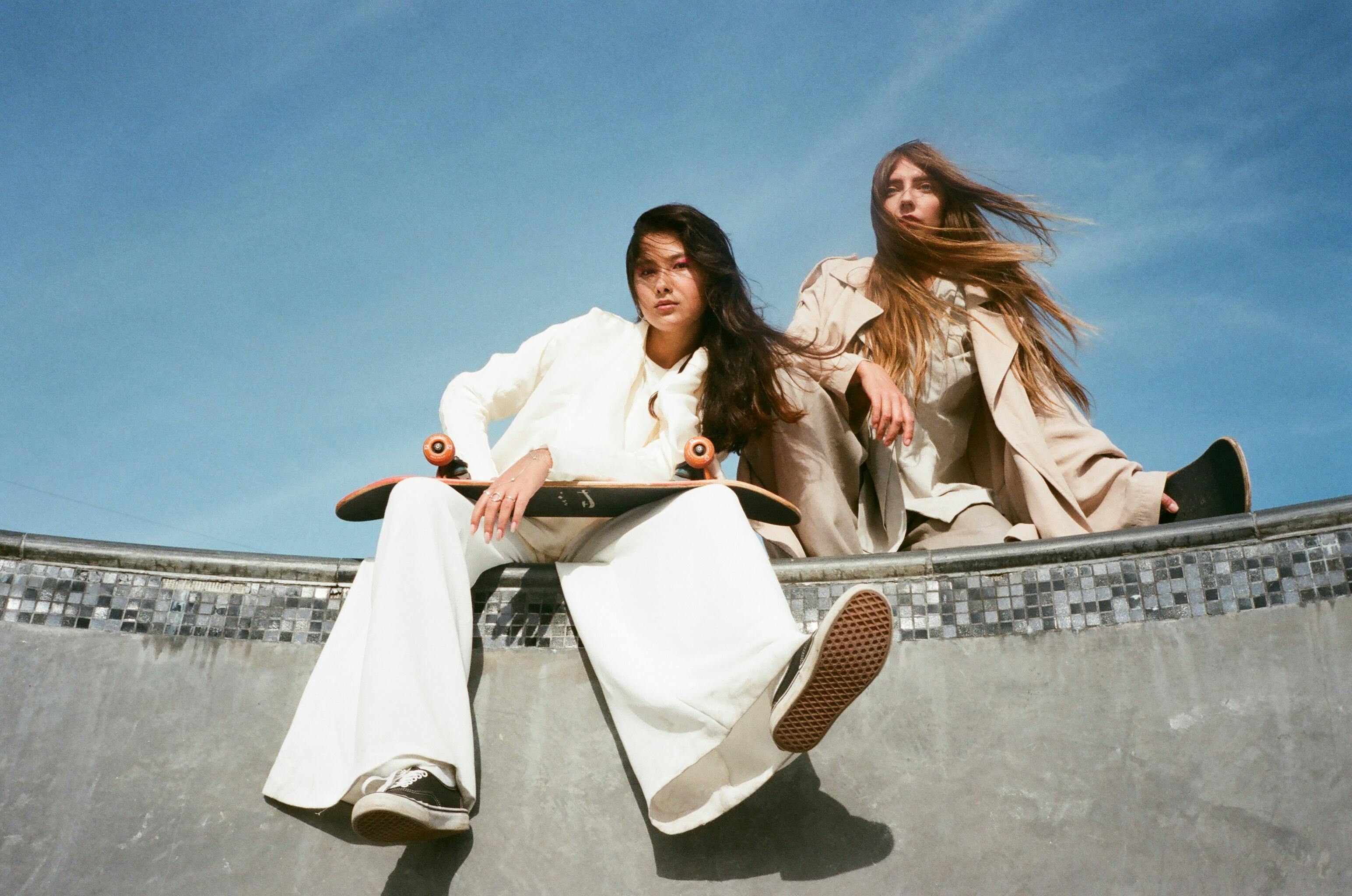 Two people with skateboards sit in a skate park under a blue sky.