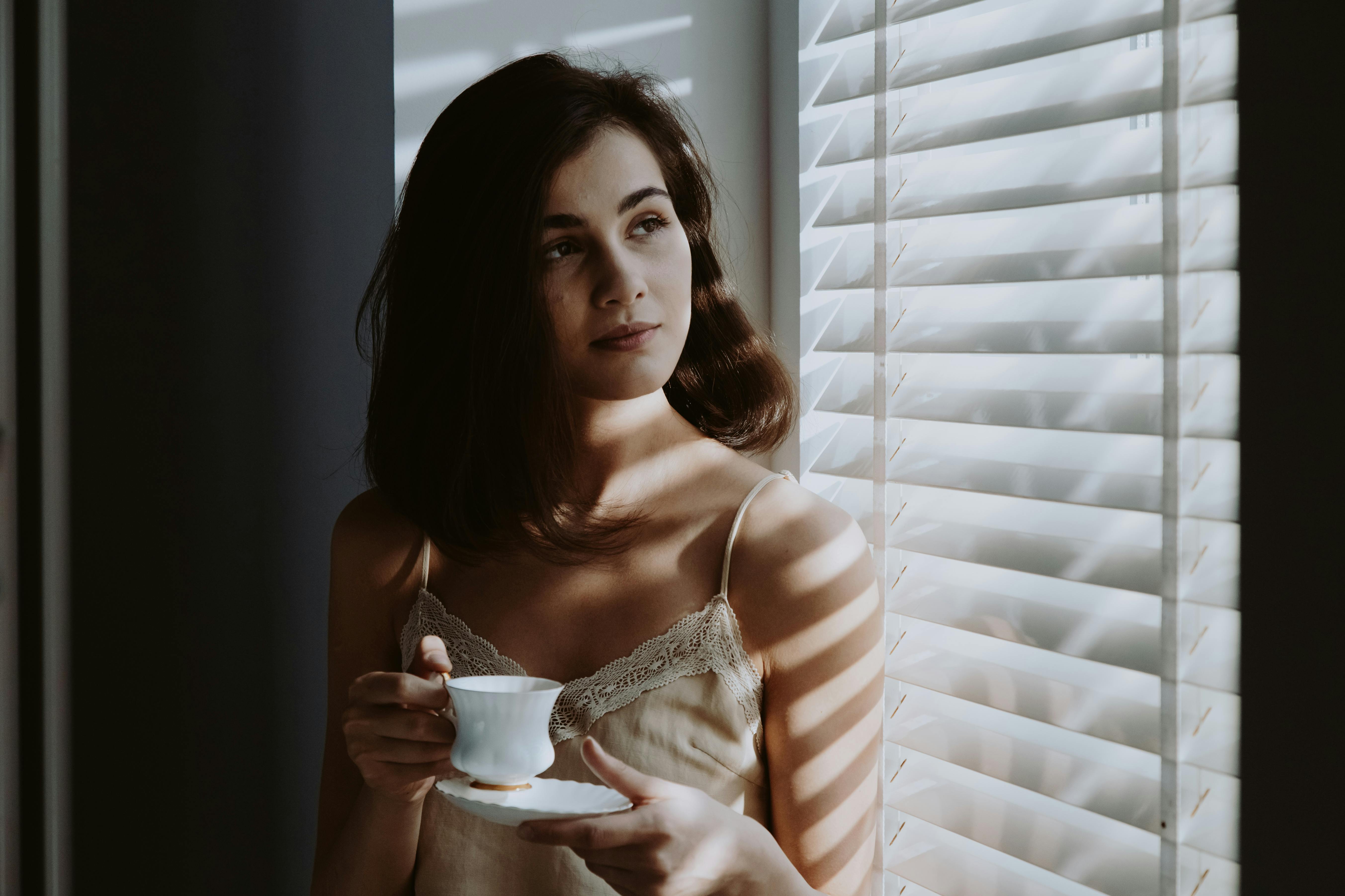 Person holding teacup, standing by window with blinds, thoughtful.