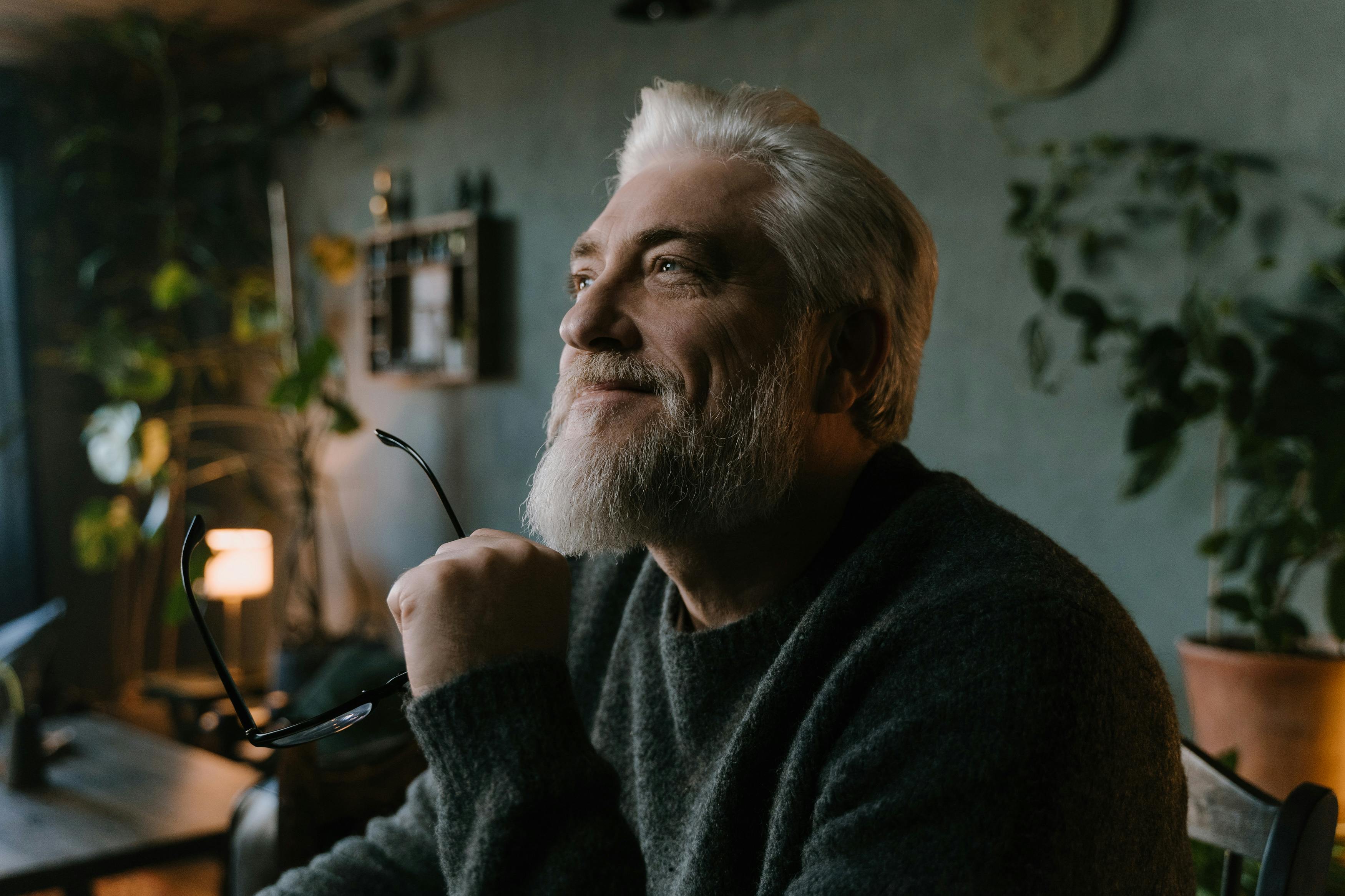 Person smiling thoughtfully indoors, holding glasses, surrounded by plants.