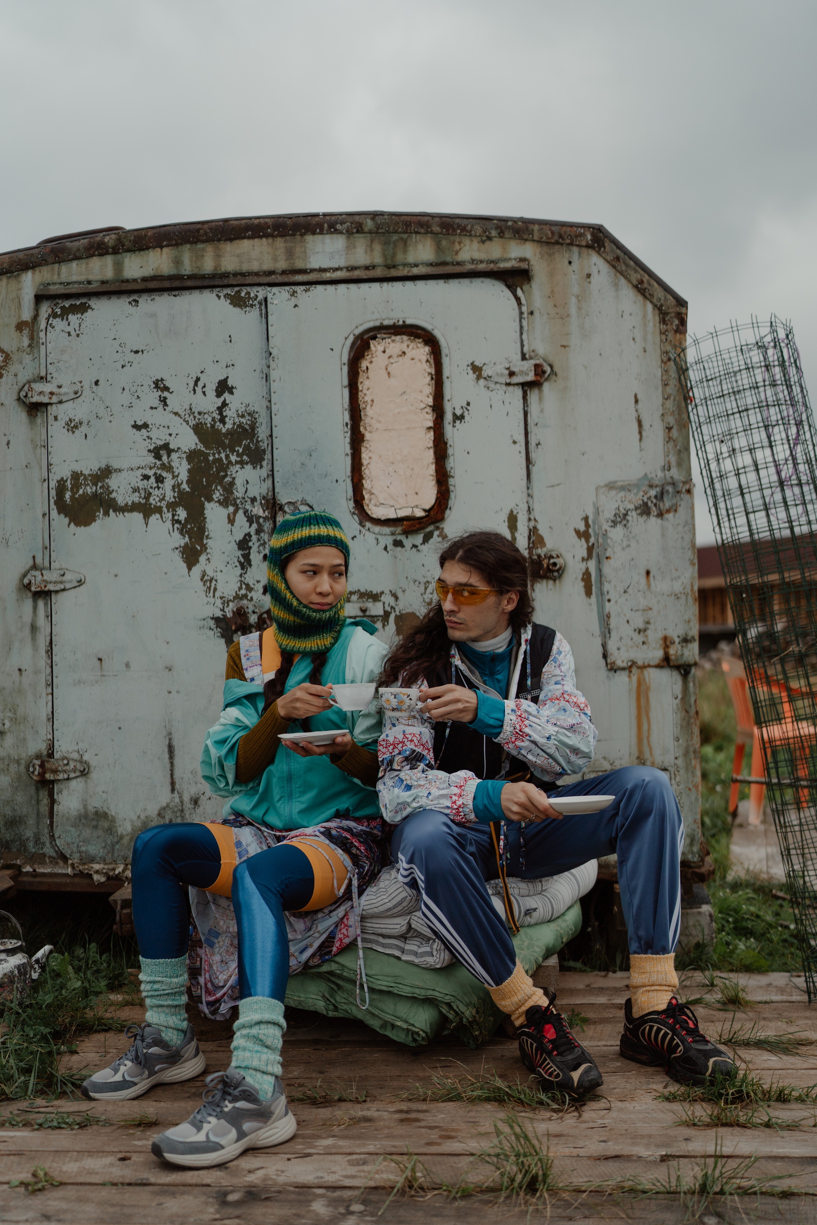 Two people in colorful outfits sit outside, drinking tea together.