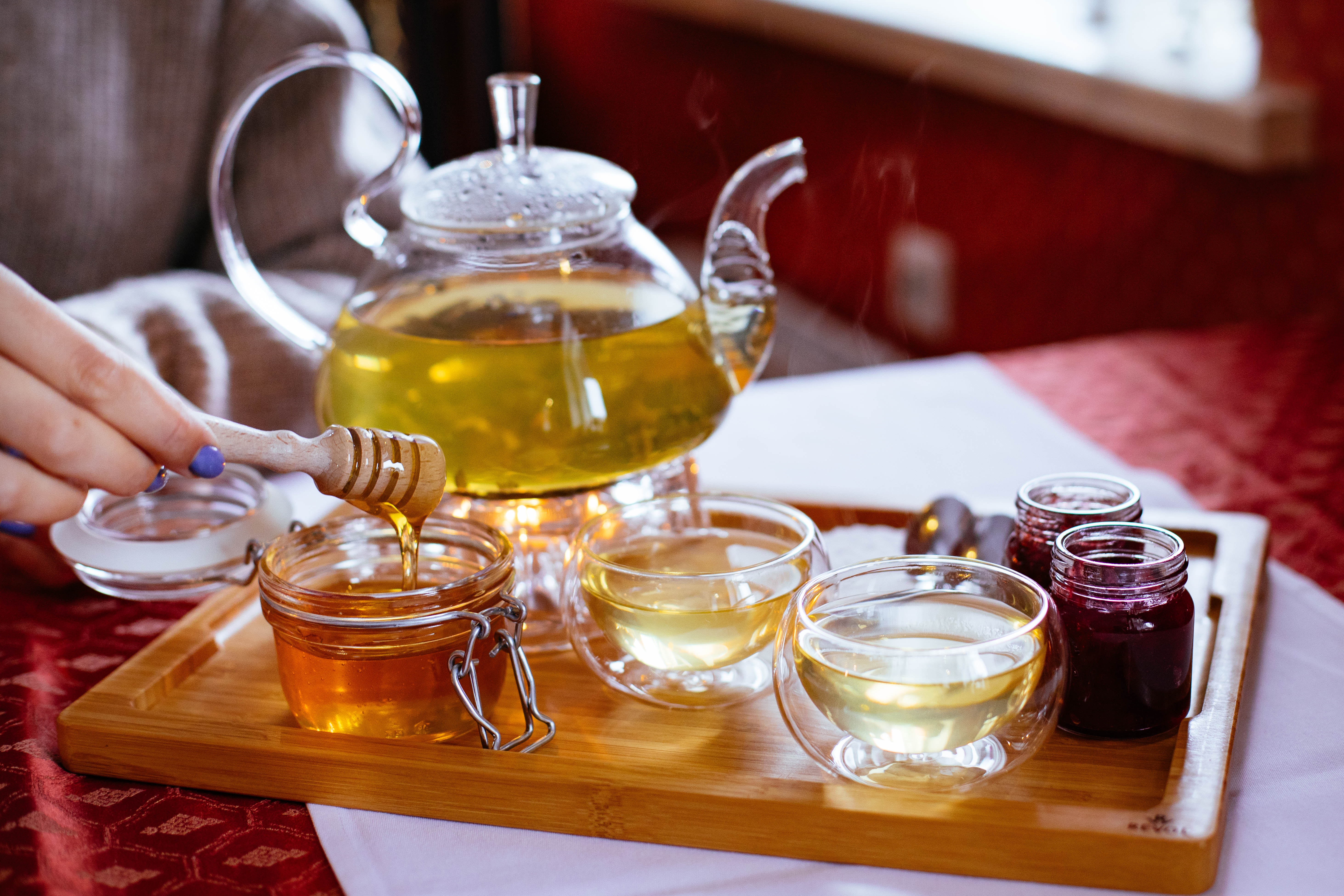 Glass teapot, tea cups, honey, and jam on wooden tray.