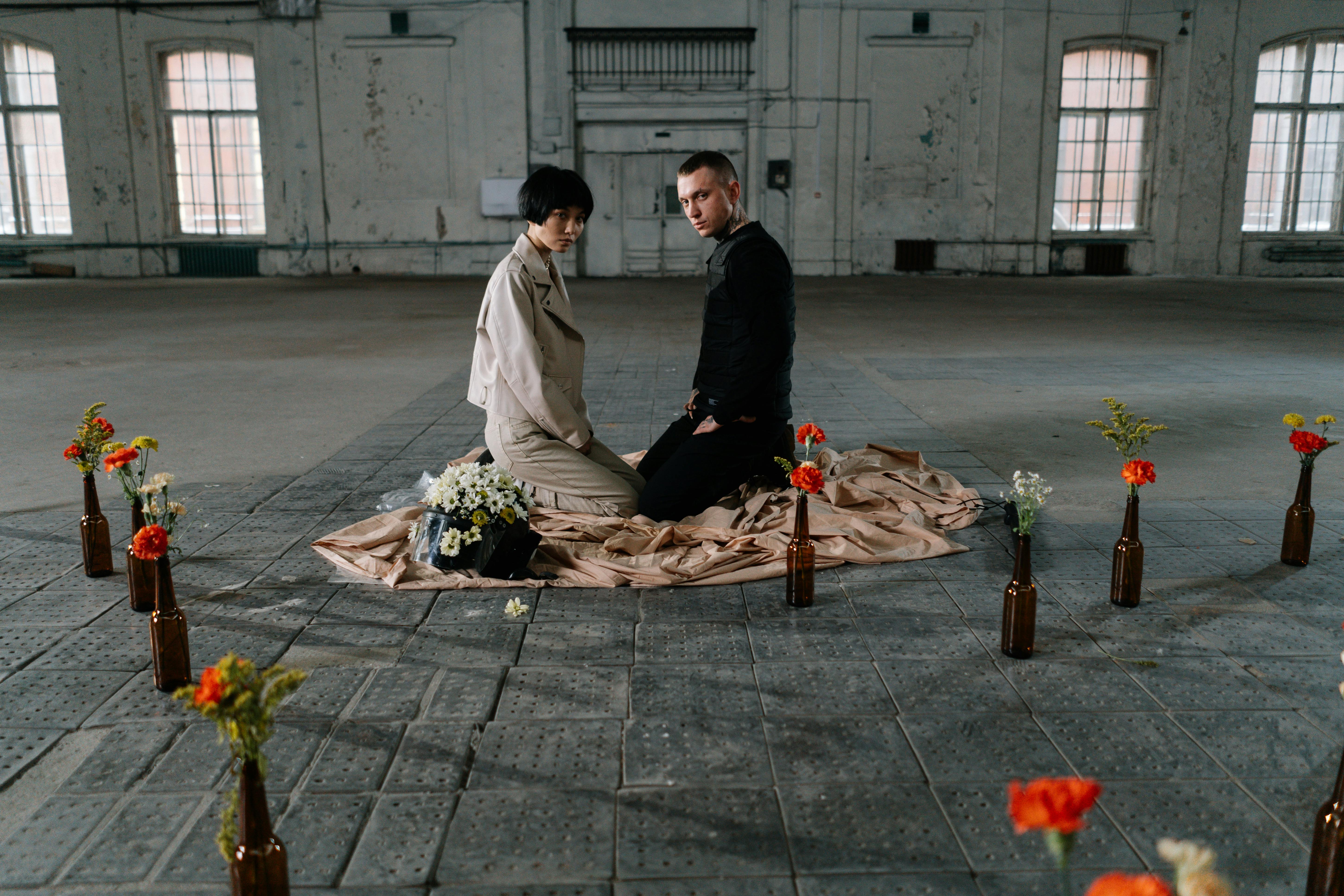 Two people kneel on fabric, surrounded by bottles with flowers.