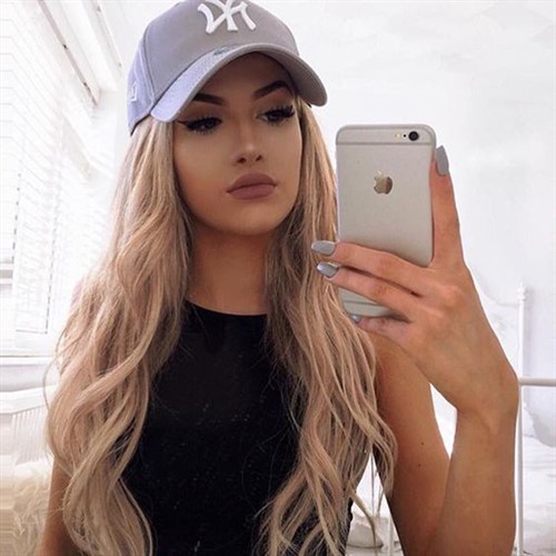 A young woman with blonde hair and a grey baseball cap takes a selfie.