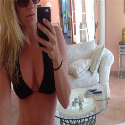 Single woman in black bikini takes a selfie in a living room.