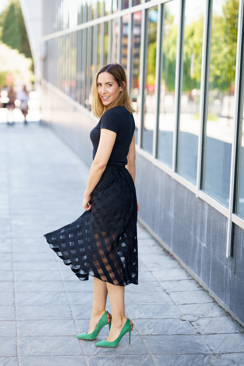 Woman in black dress and green heels twirls outside modern building.