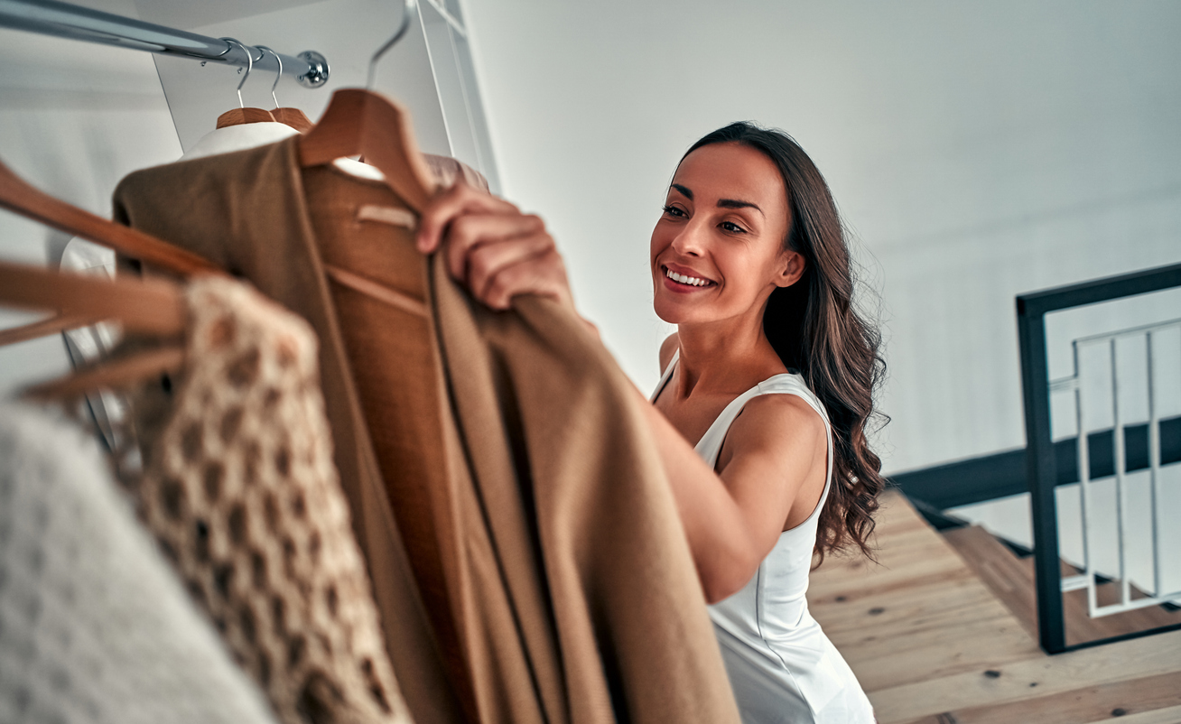 Smiling woman in white dress selecting a brown coat from a closet.