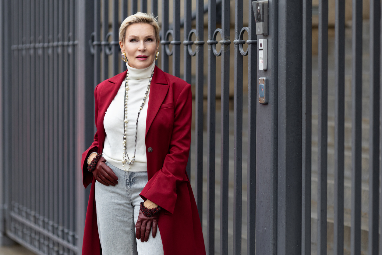 Stylish blonde woman in a red coat and gloves stands by a gate.