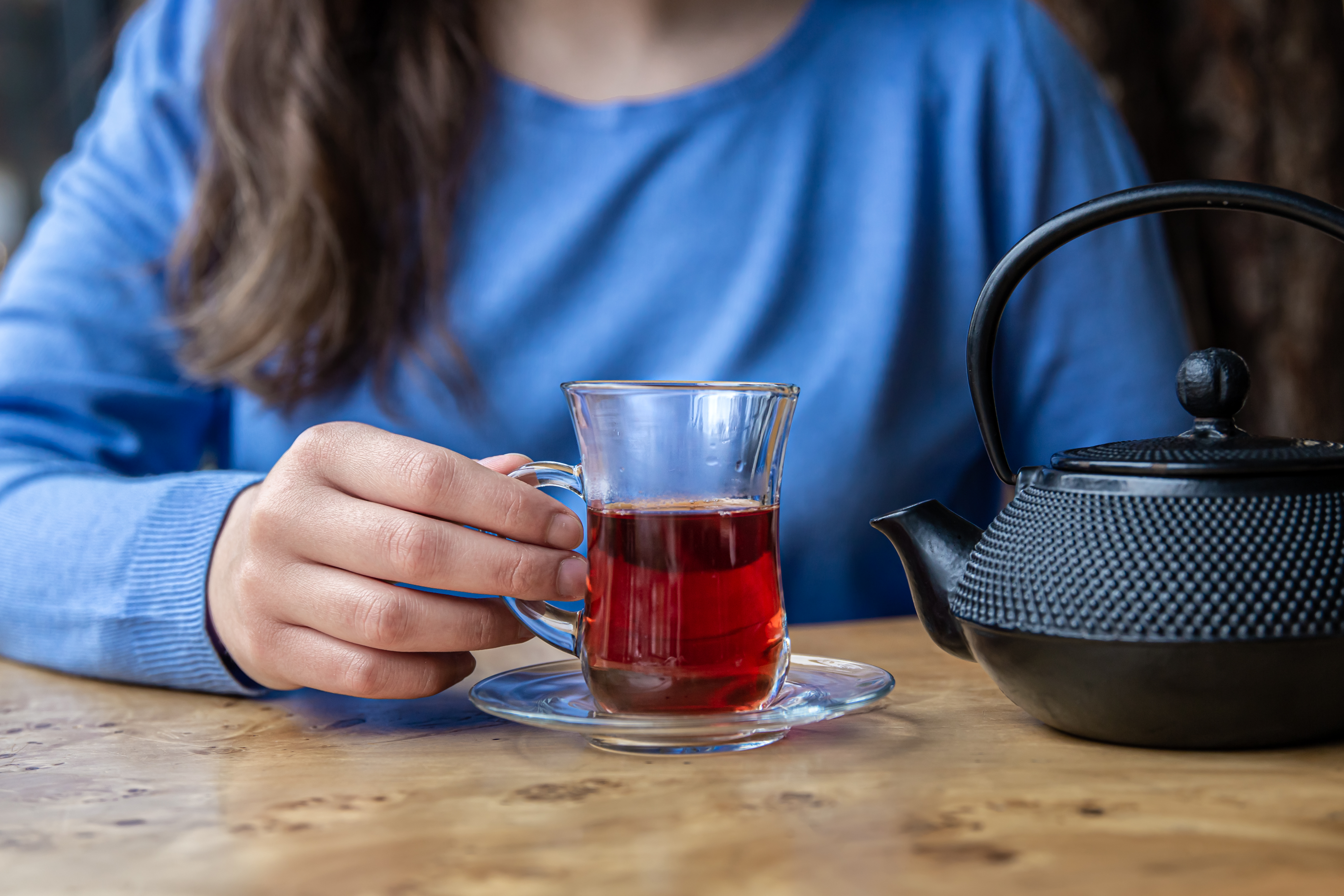 Una persona con un suéter azul sostiene un vaso de té de jamaica junto a una tetera negra.