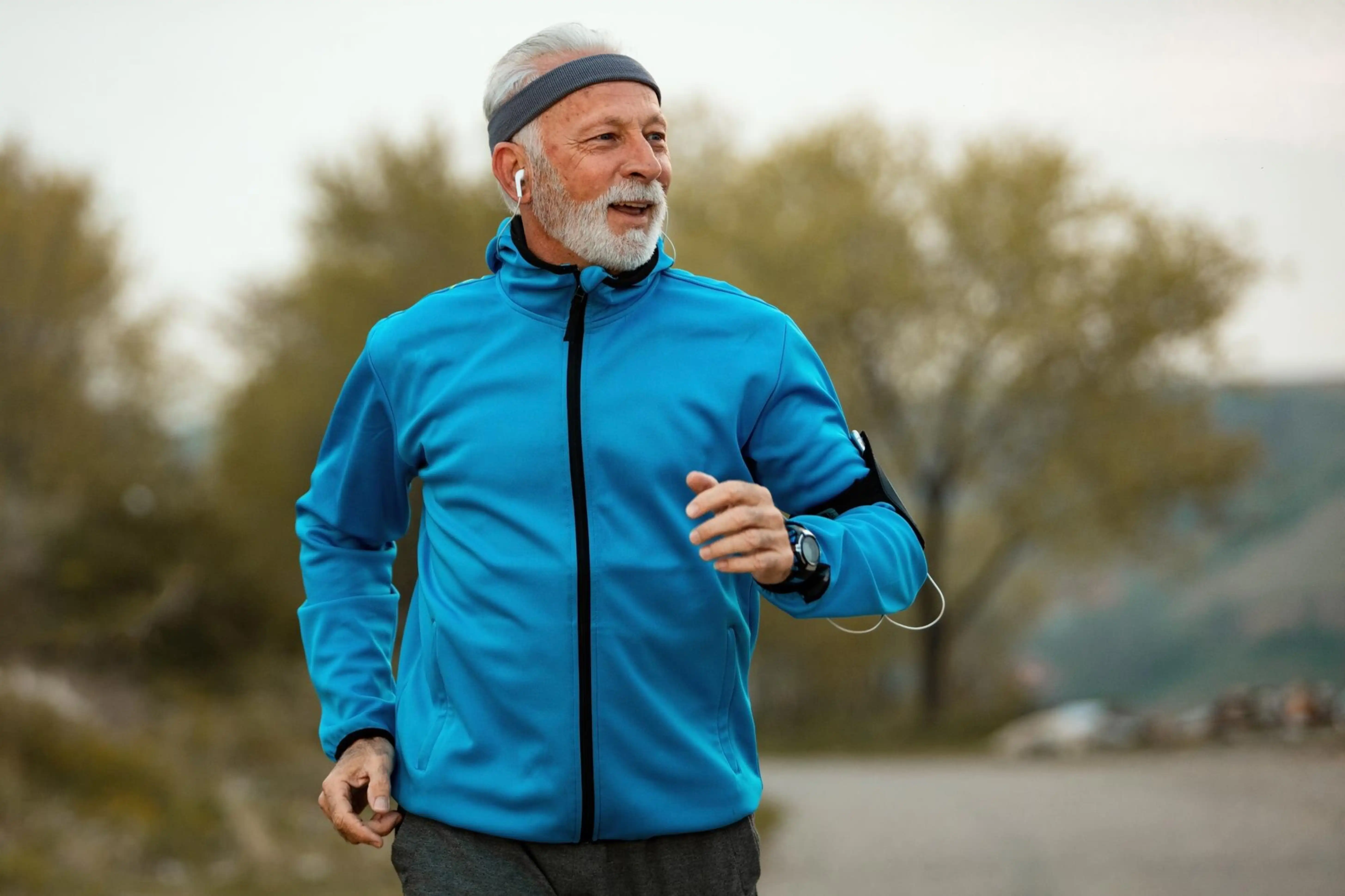 Un anciano haciendo jogging al aire libre, con auriculares y una chaqueta azul.