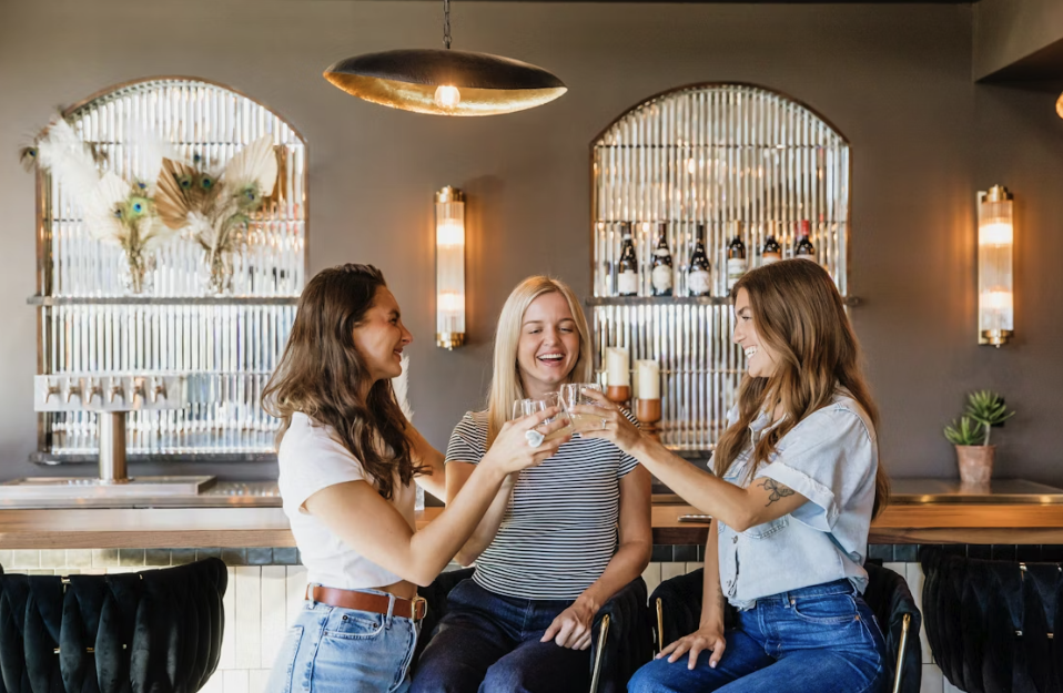 Three women clinking glasses and laughing at a bar.