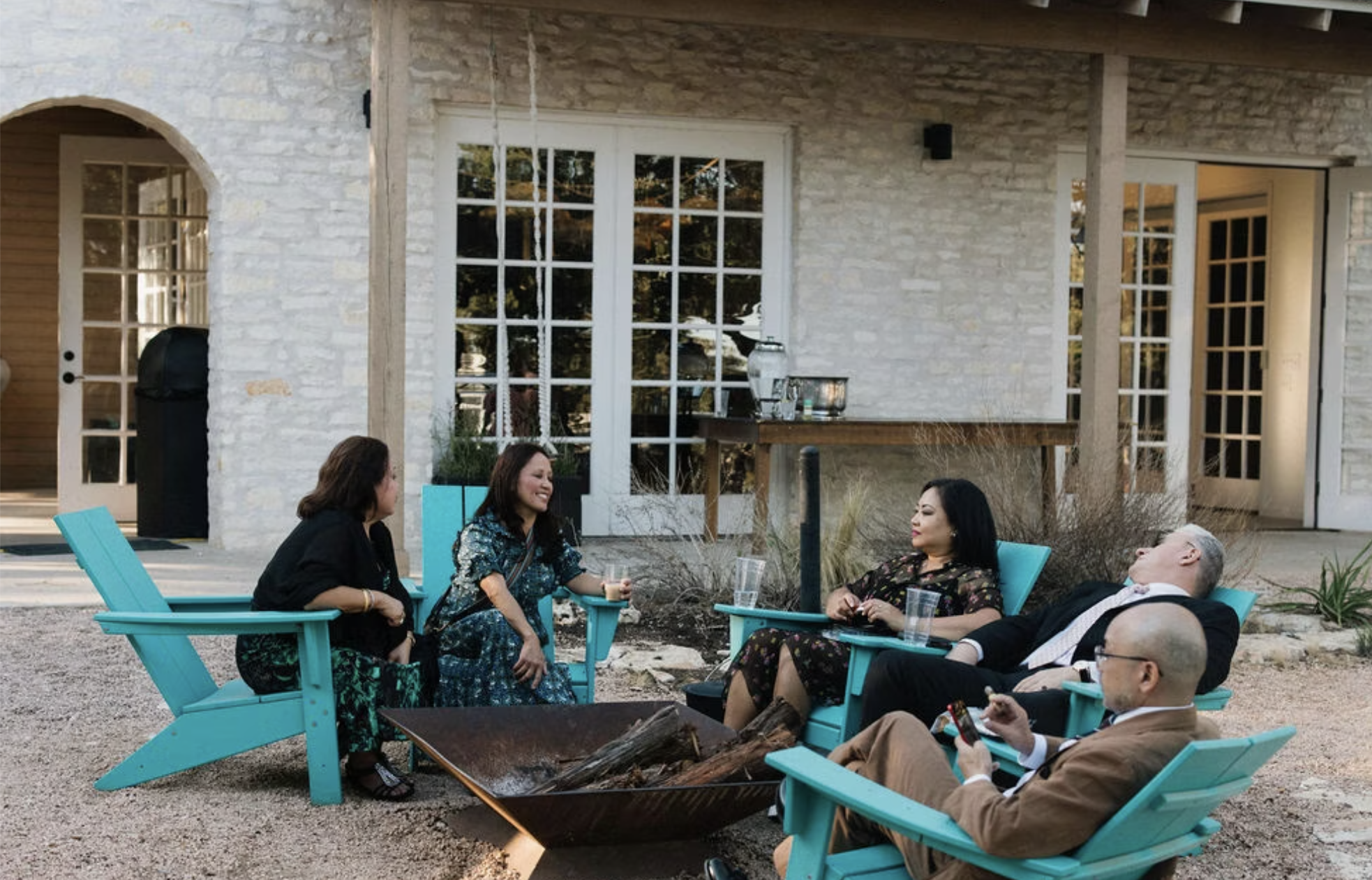 People relaxing in Adirondack chairs around a fire pit outside a stone building.